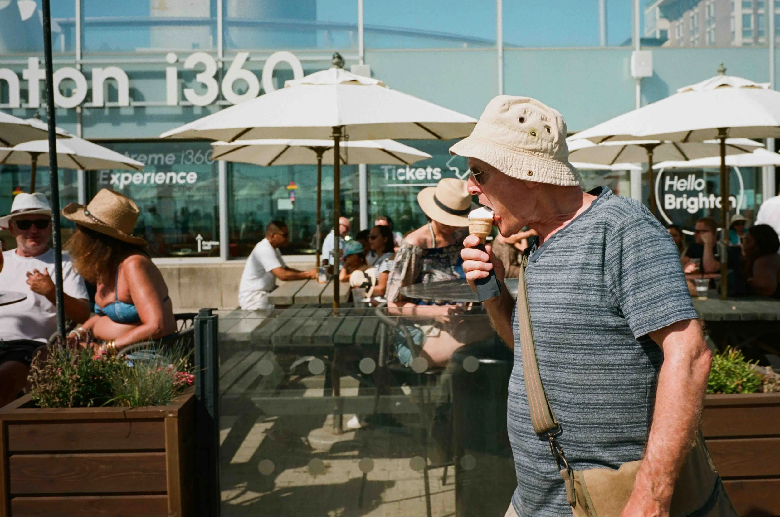 A man wearing a bucket hat and sunglasses eating ice cream outdoors at a social gathering with people under umbrellas.
