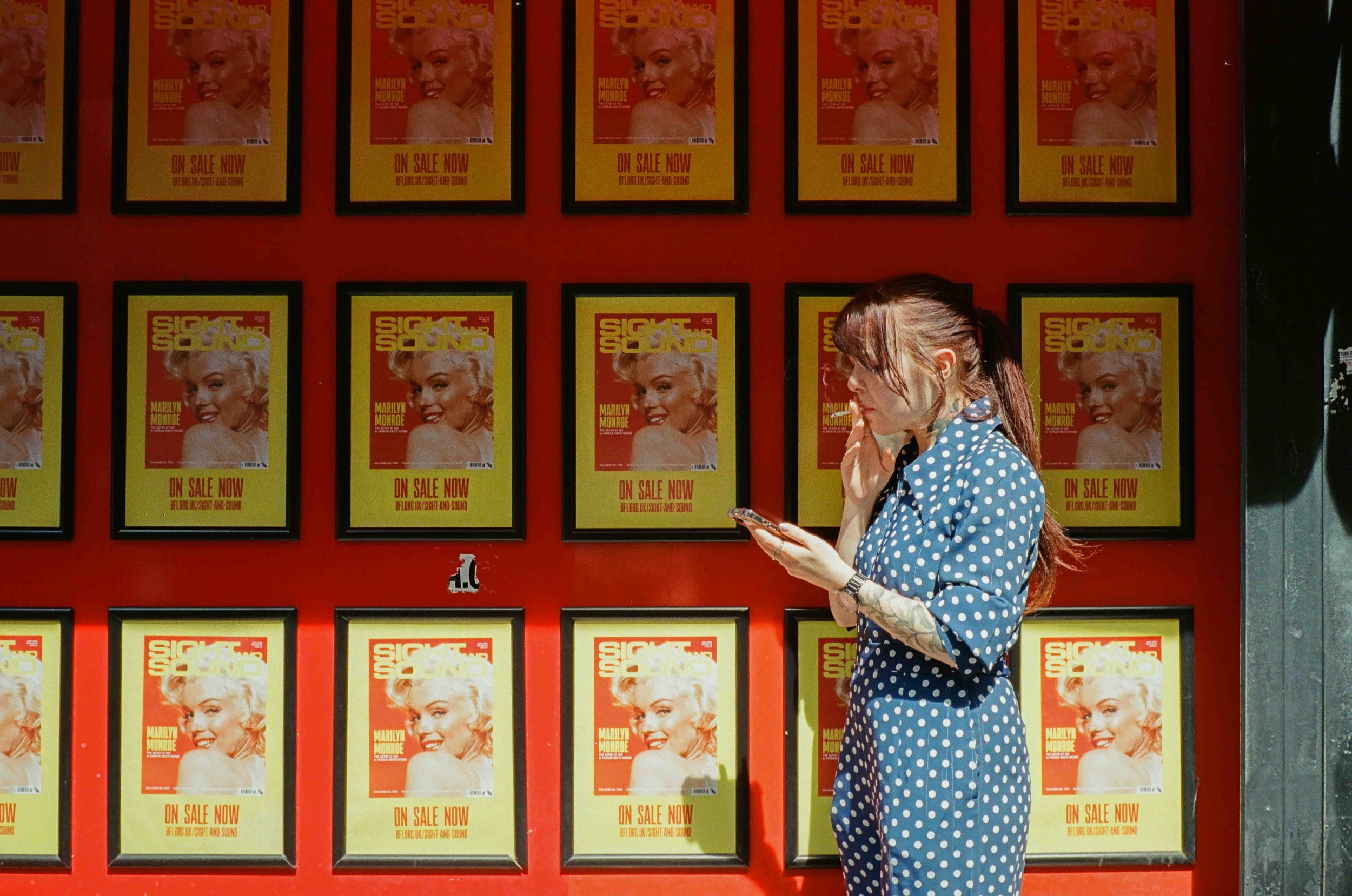 A woman with tattoos in a blue polka dot dress looking at her phone in front of a wall decorated with posters of Marilyn Monroe advertising an upcoming show.