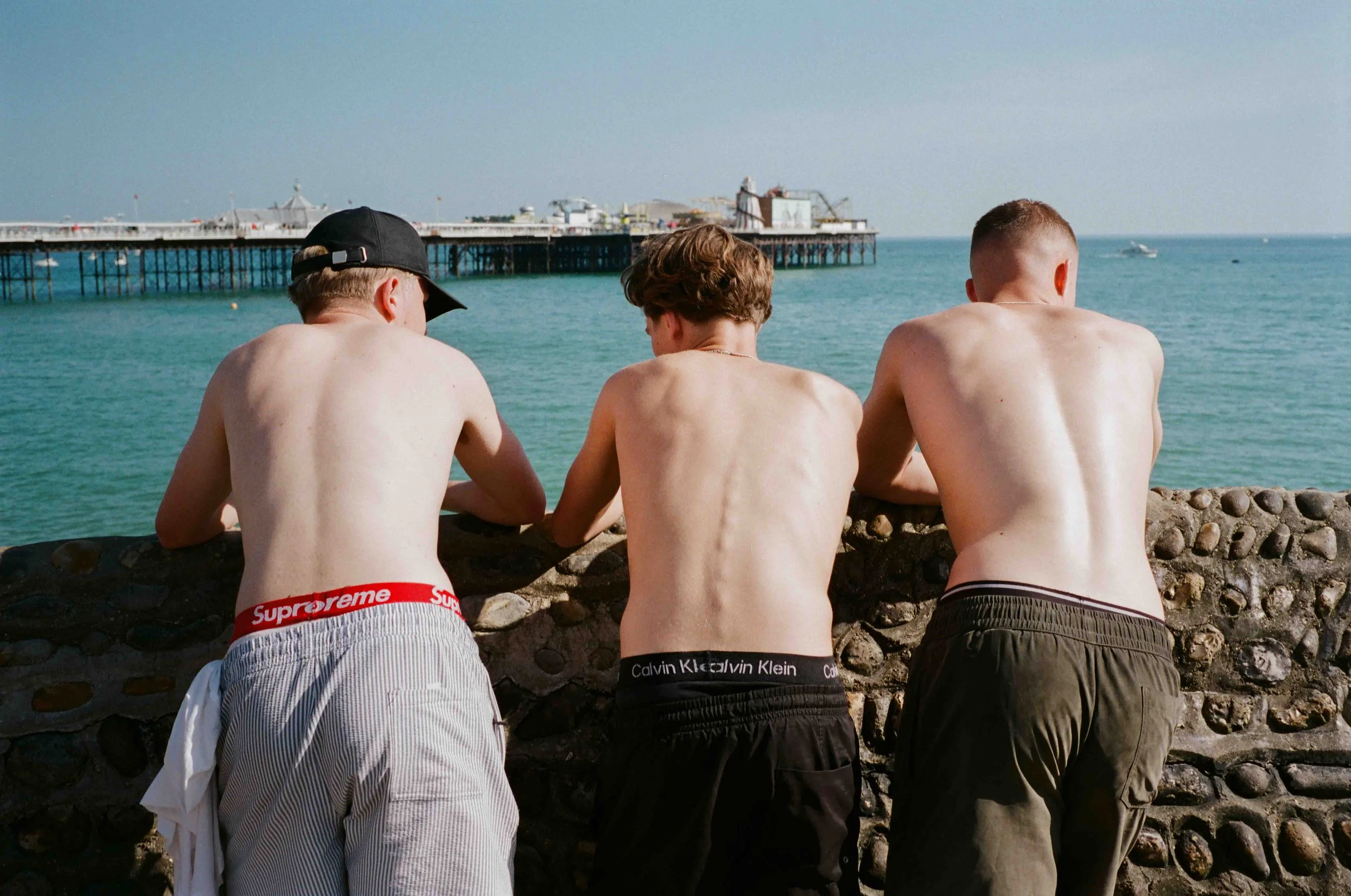 Three shirtless young men sitting on a stone ledge by the ocean, facing away, with a pier in the background.