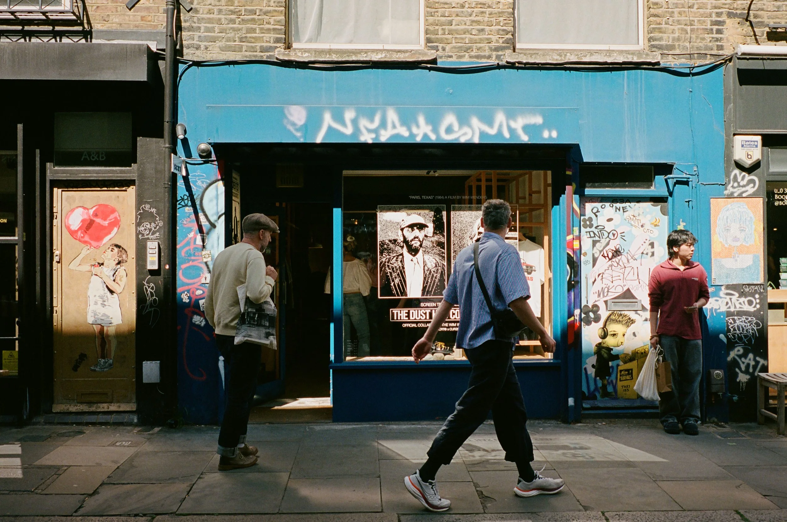 People walking on a city sidewalk in front of a building with graffiti and street art, including a large portrait of a man with glasses and a hat, and a poster of a woman with food, under a blue awning.