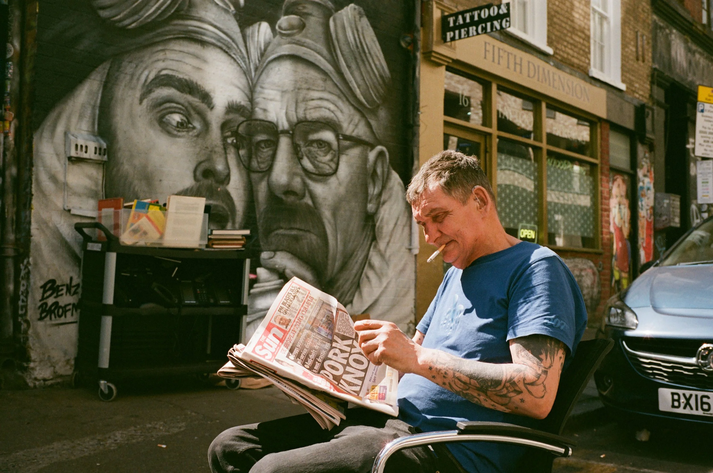 A man with tattoos on his arm, wearing a blue shirt, sitting outside and reading a newspaper, with a cigarette in his mouth. Behind him is a mural of a woman with glasses, and various storefronts and posters are visible in the background.