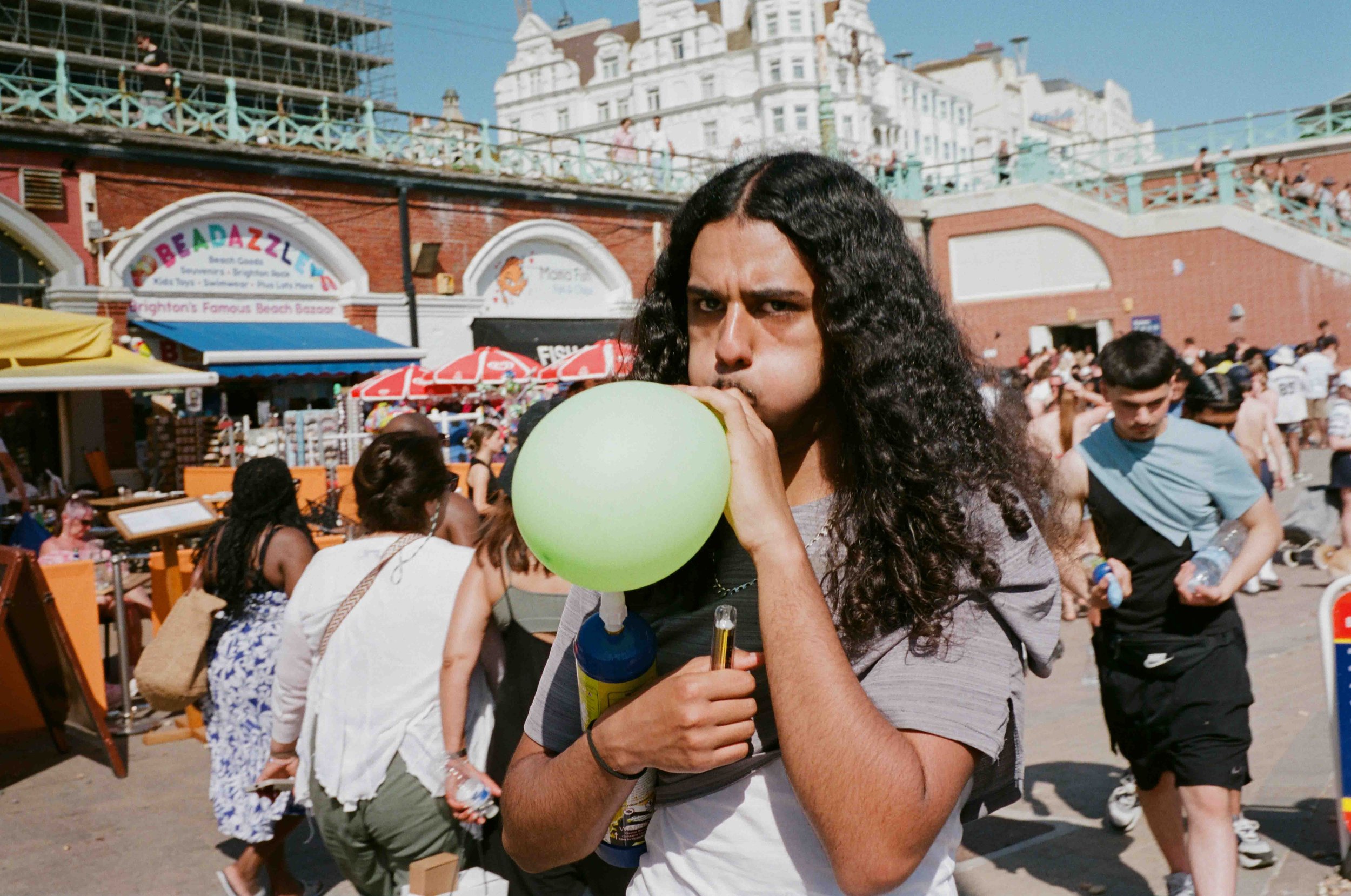 A young person with long curly dark hair blowing up a green balloon at a crowded outdoor market or fair, with colorful stalls and people in the background. Buildings and a bridge are visible in the distance.
