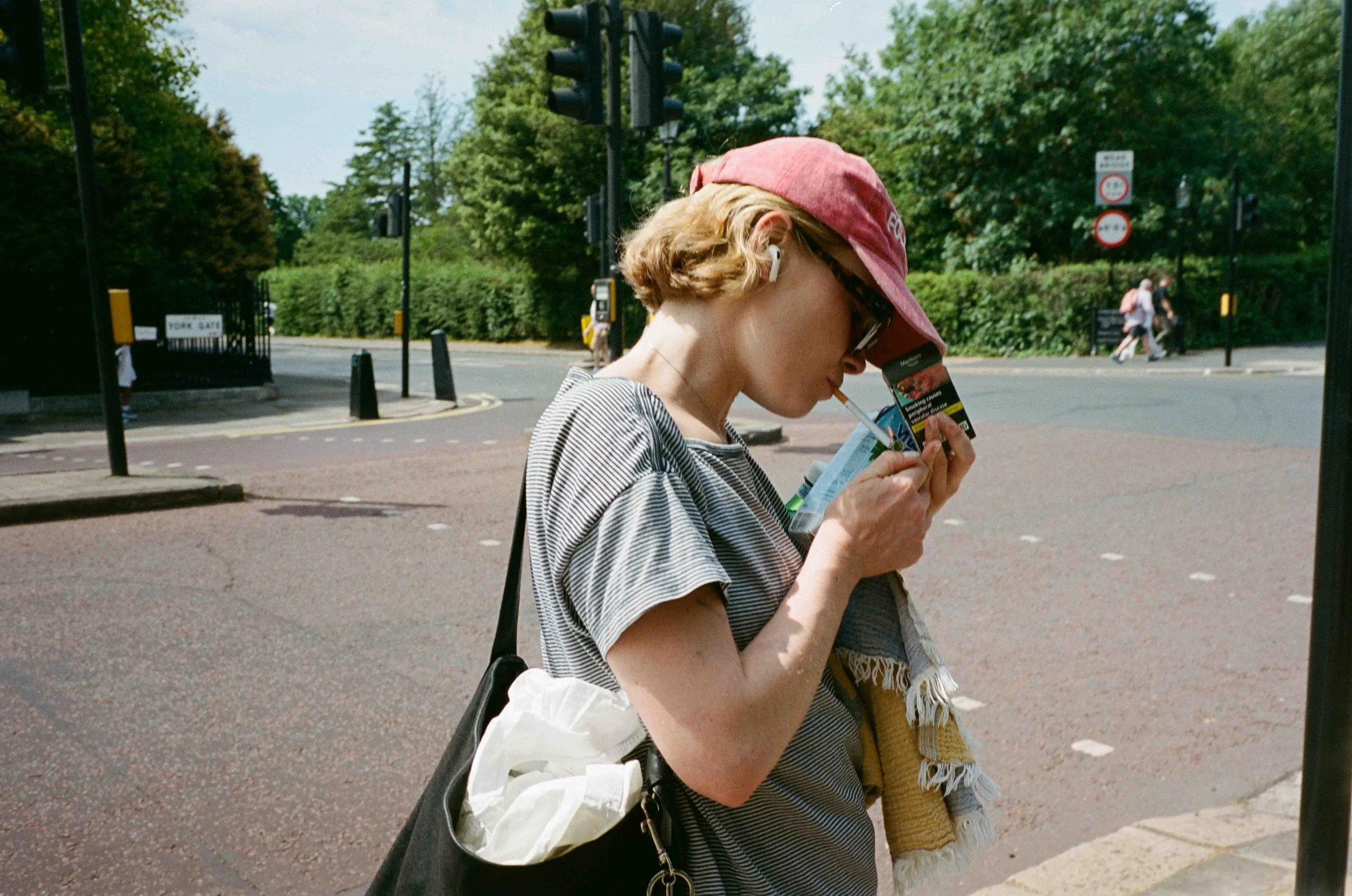 A young woman with short curly blonde hair, wearing a pink cap and striped shirt, is standing outdoors on a city street. She has AirPods in her ears and is looking down at her phone while holding a cigarette and a pack of cards. She carries a black h
