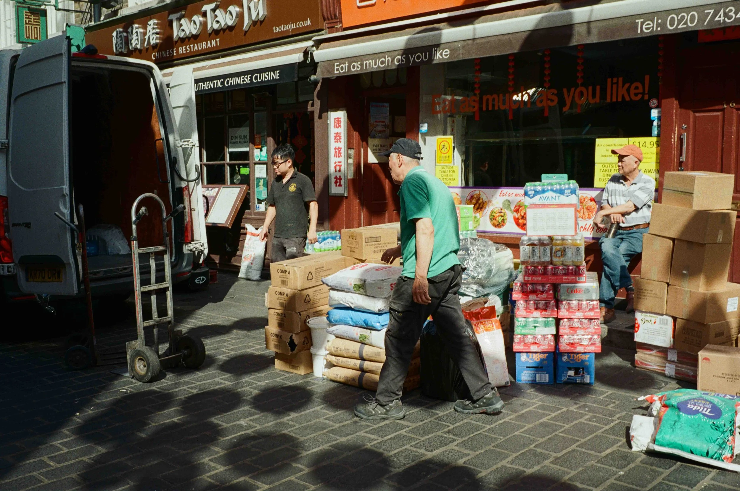 People unloading goods and supplies outside a Chinese restaurant named Taotaoju, with stacks of boxes, bottled drinks, and bags of food on the street.