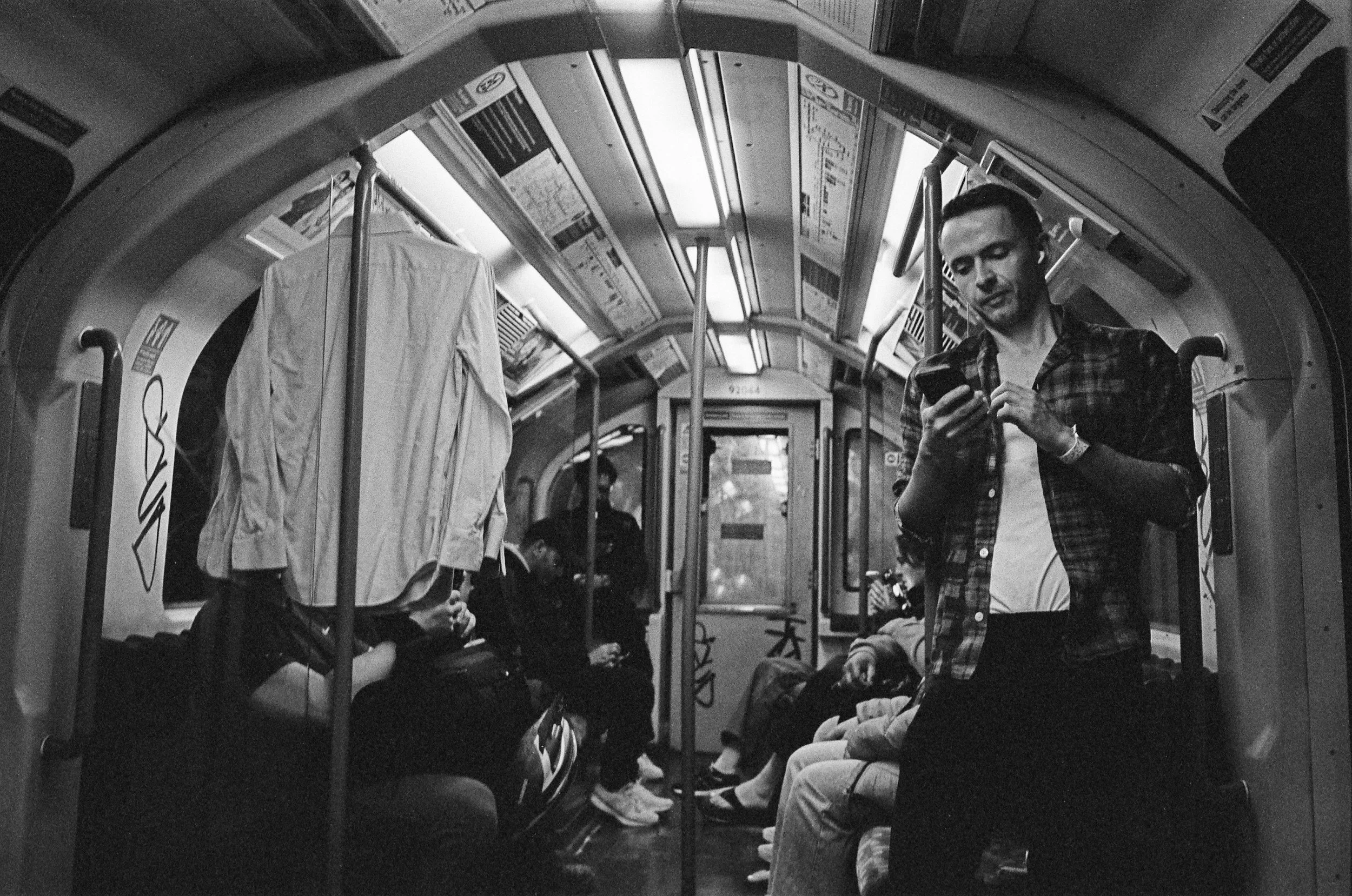 A black and white photo inside a subway or train car showing a man standing and looking at his phone. Other passengers are seated, some with headphones and bags, while a large item covered with a cloth is on the left side of the image. The overhead s