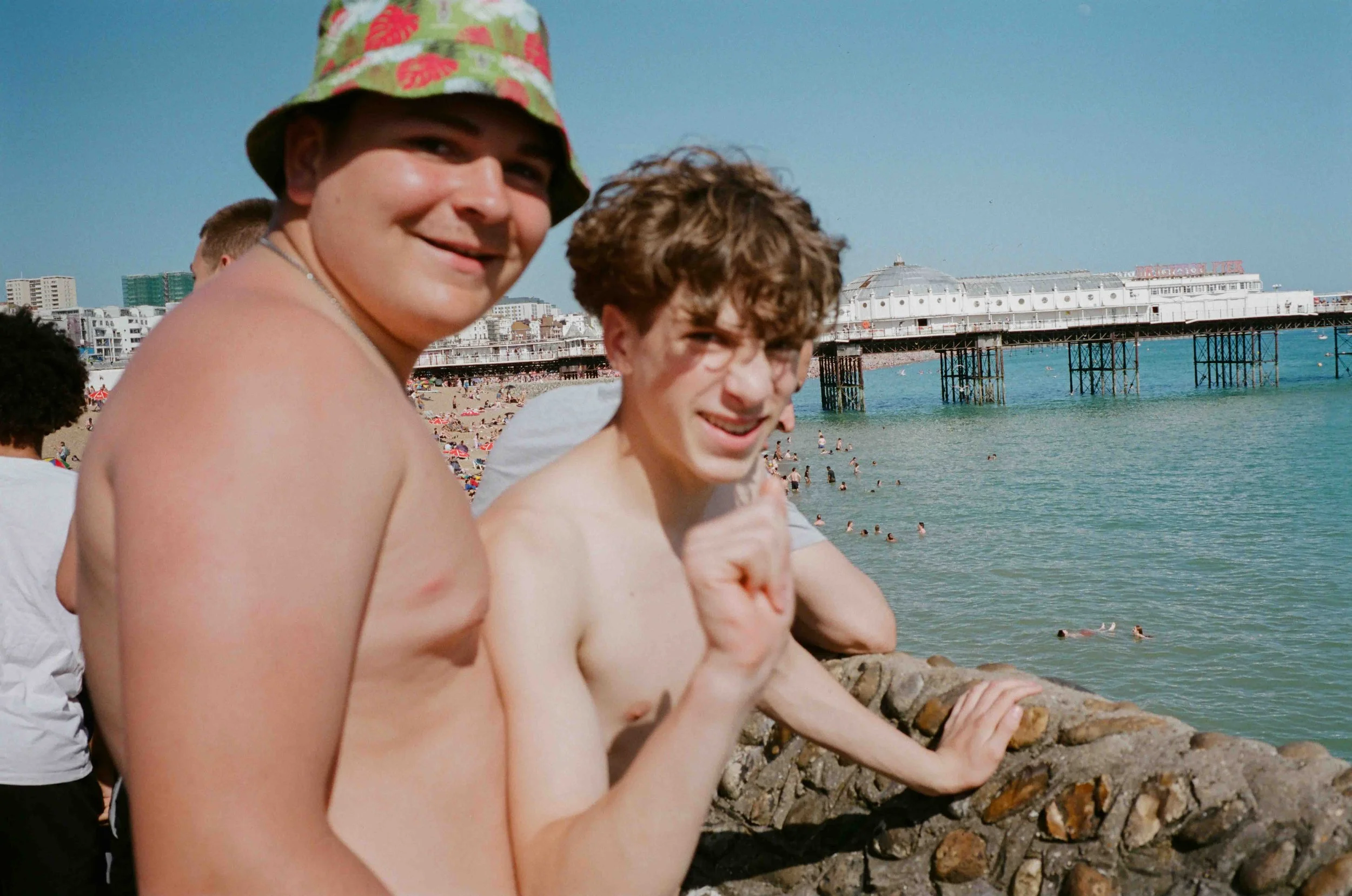 Young people sitting on rocks at the beach, smiling and enjoying a sunny day with the ocean, a pier, and colorful beach umbrellas in the background.