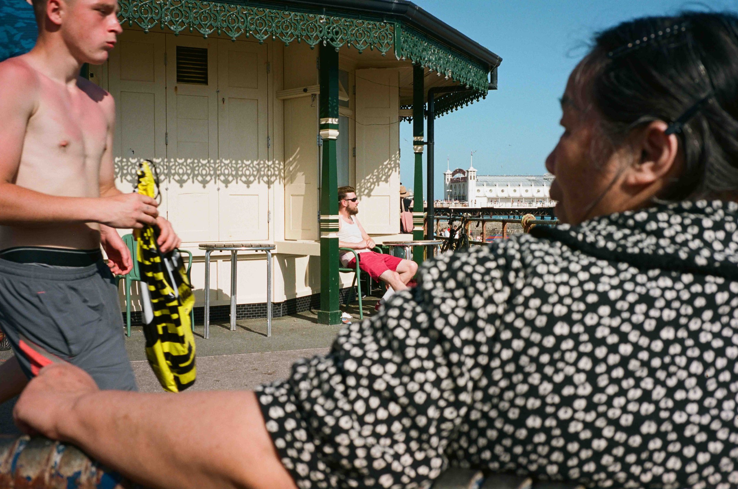 A shirtless man holding a yellow and black item, possibly a towel or clothing, on a boardwalk or pier with a Victorian-style building and blue sky in the background. A woman with dark hair and glasses, wearing a polka-dot top, is partially visible in