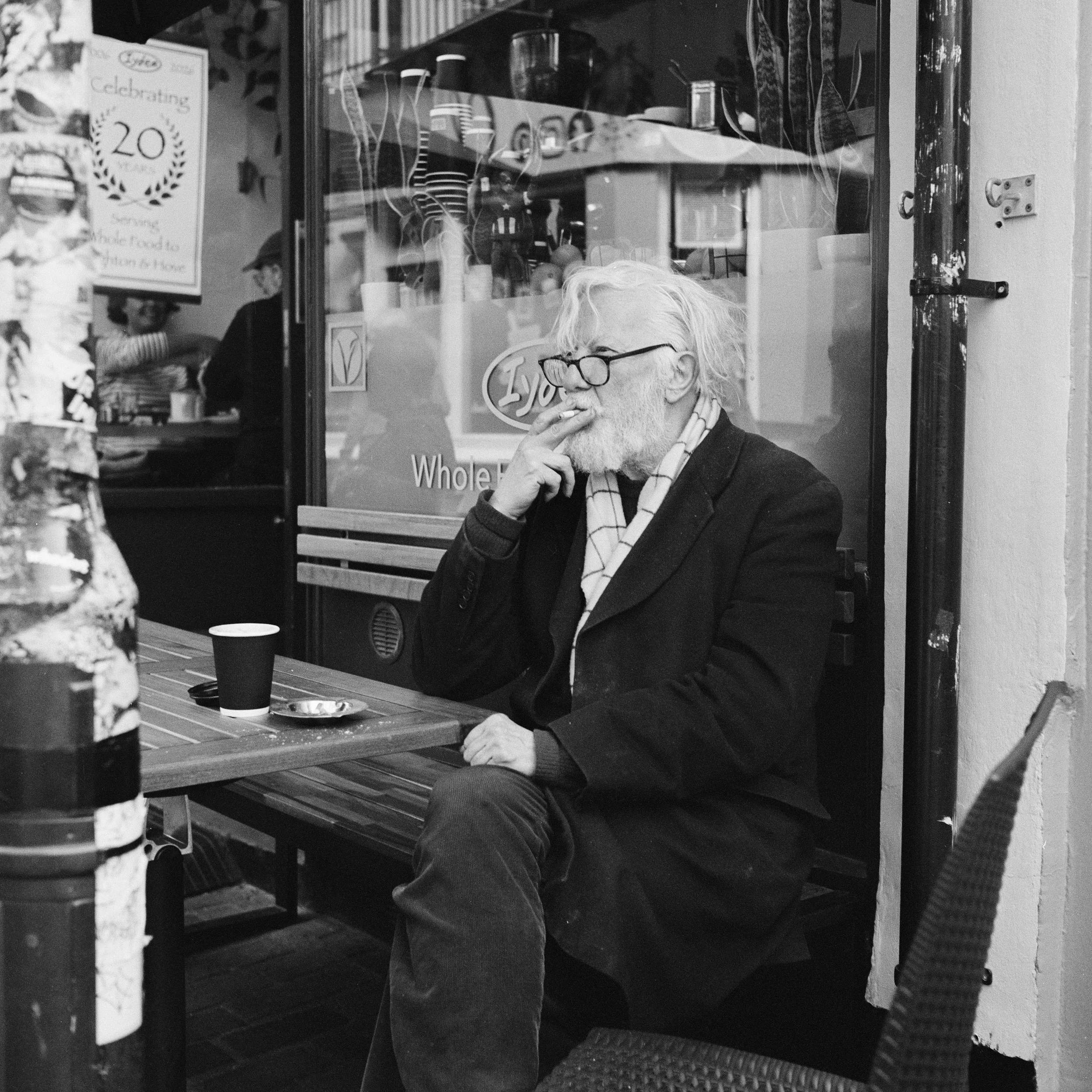 An elderly man with white hair and beard, wearing glasses and a striped scarf, sitting at an outdoor table and smoking a cigarette inside a cafe or restaurant.