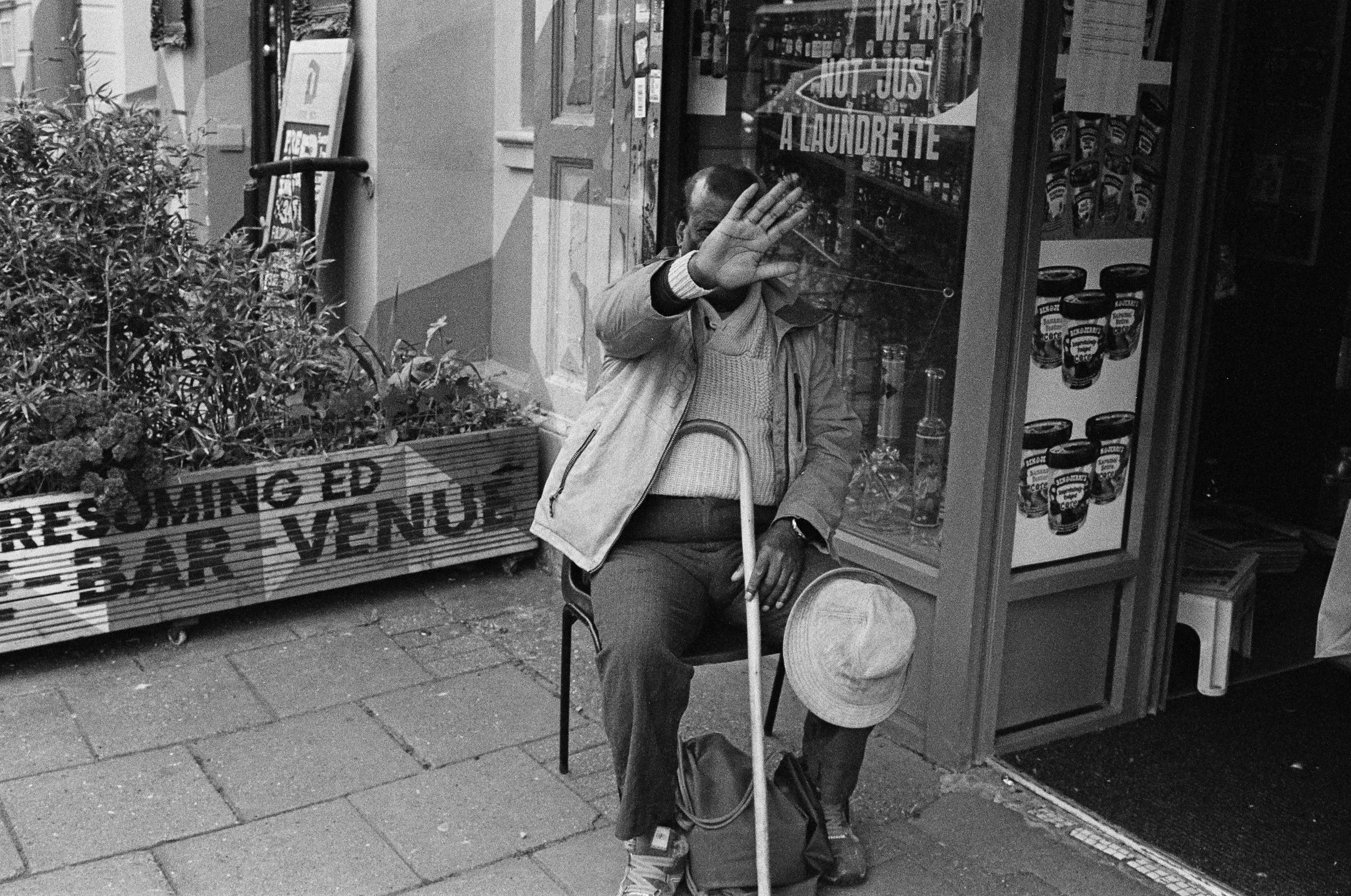 A man sitting on a folding chair outside a store, raising one hand to block the camera, with a shopping bag hanging from his arm, in front of a window displaying various products.