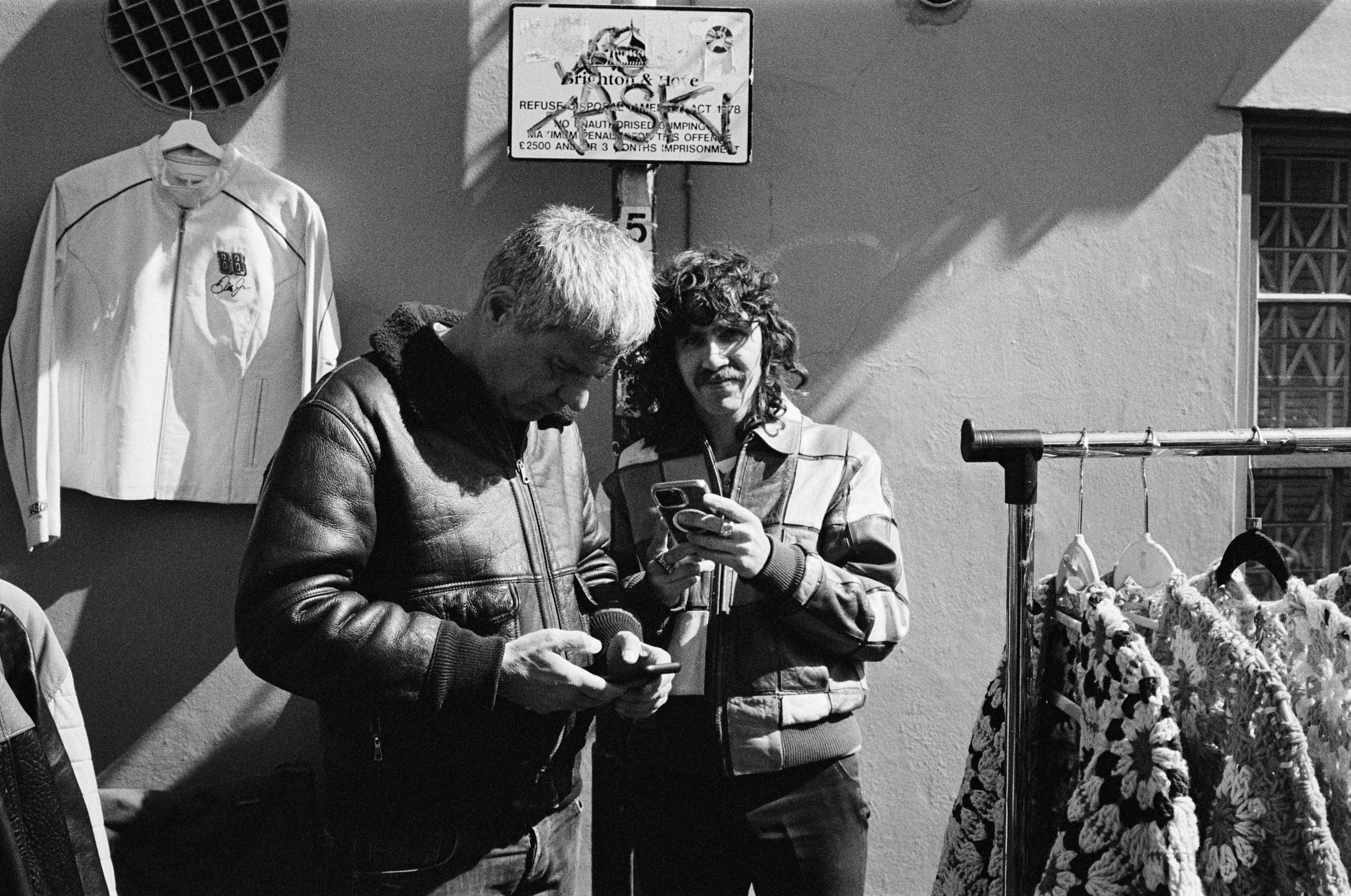 Two men are standing side by side on a sidewalk next to a wall. One is wearing a leather jacket, and the other is wearing a jacket with horizontal stripes. The man on the right appears to be looking at a phone. Behind them, there is a clothes rack wi