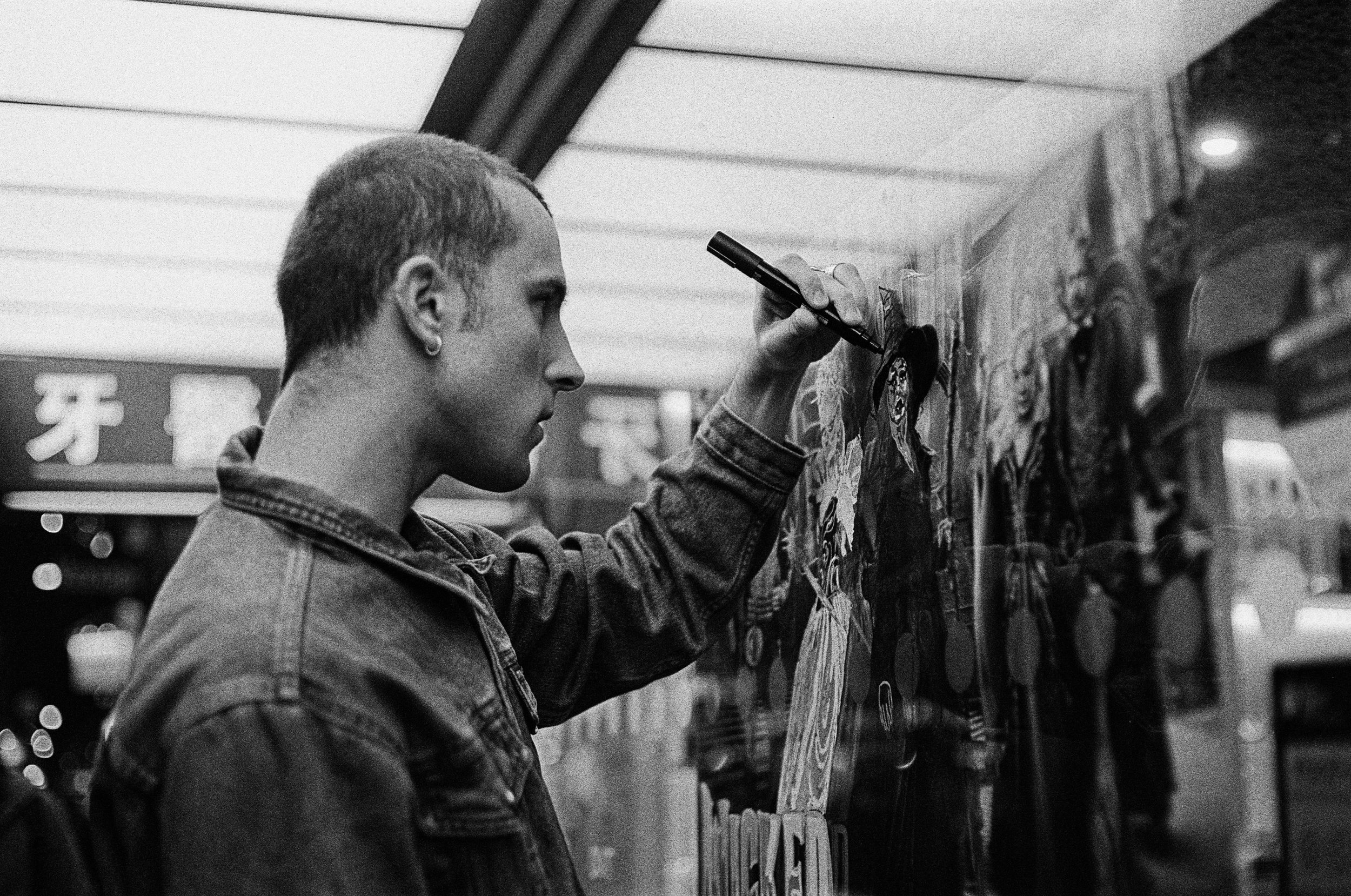 A person with short hair and a denim jacket is drawing or writing on a glass window with a marker, next to colorful posters or artwork inside a store or shop in an urban area at night.