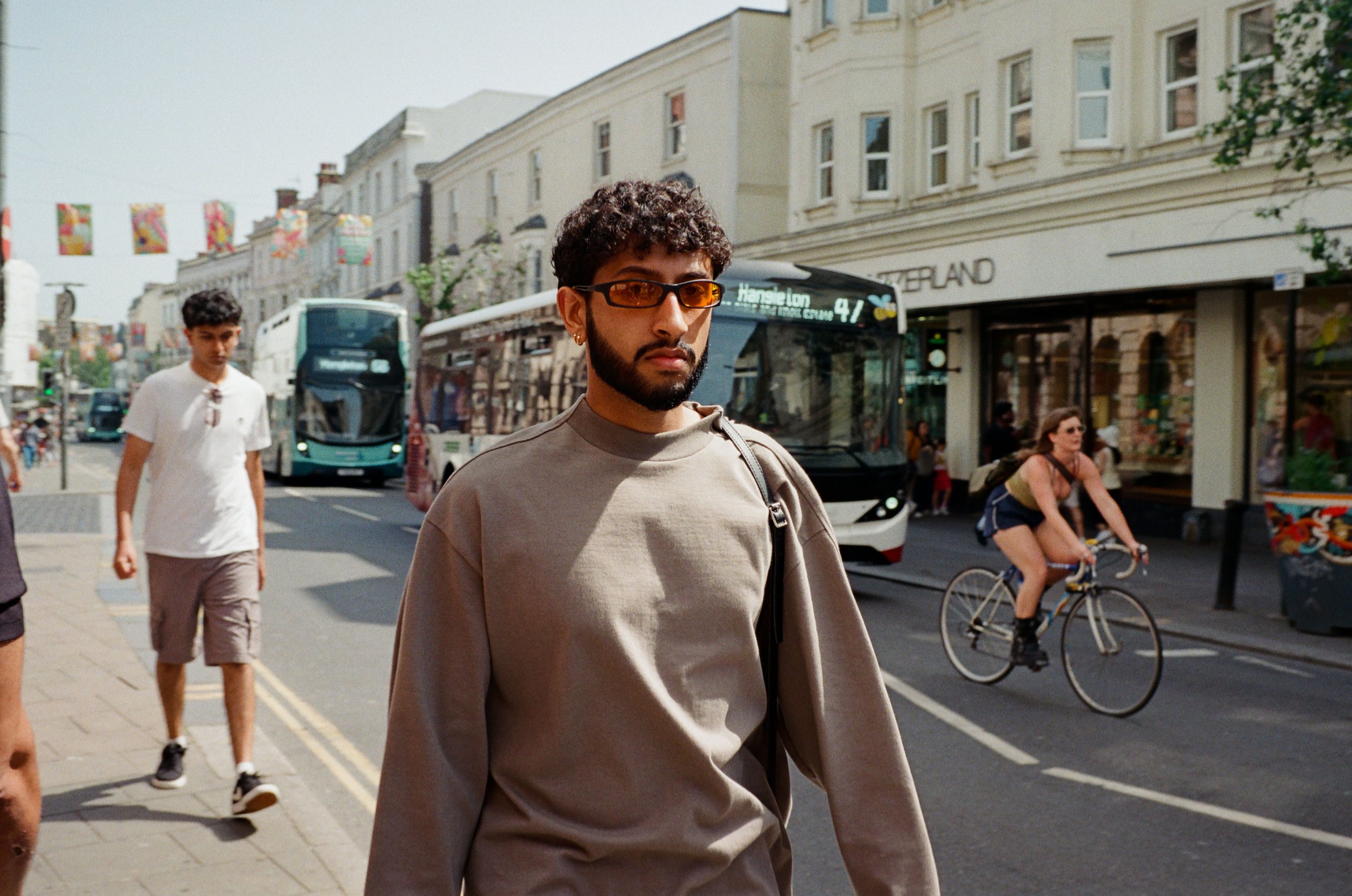 A young man with curly hair and glasses walking on a city street, with a bus, pedestrians, a cyclist, and buildings in the background.