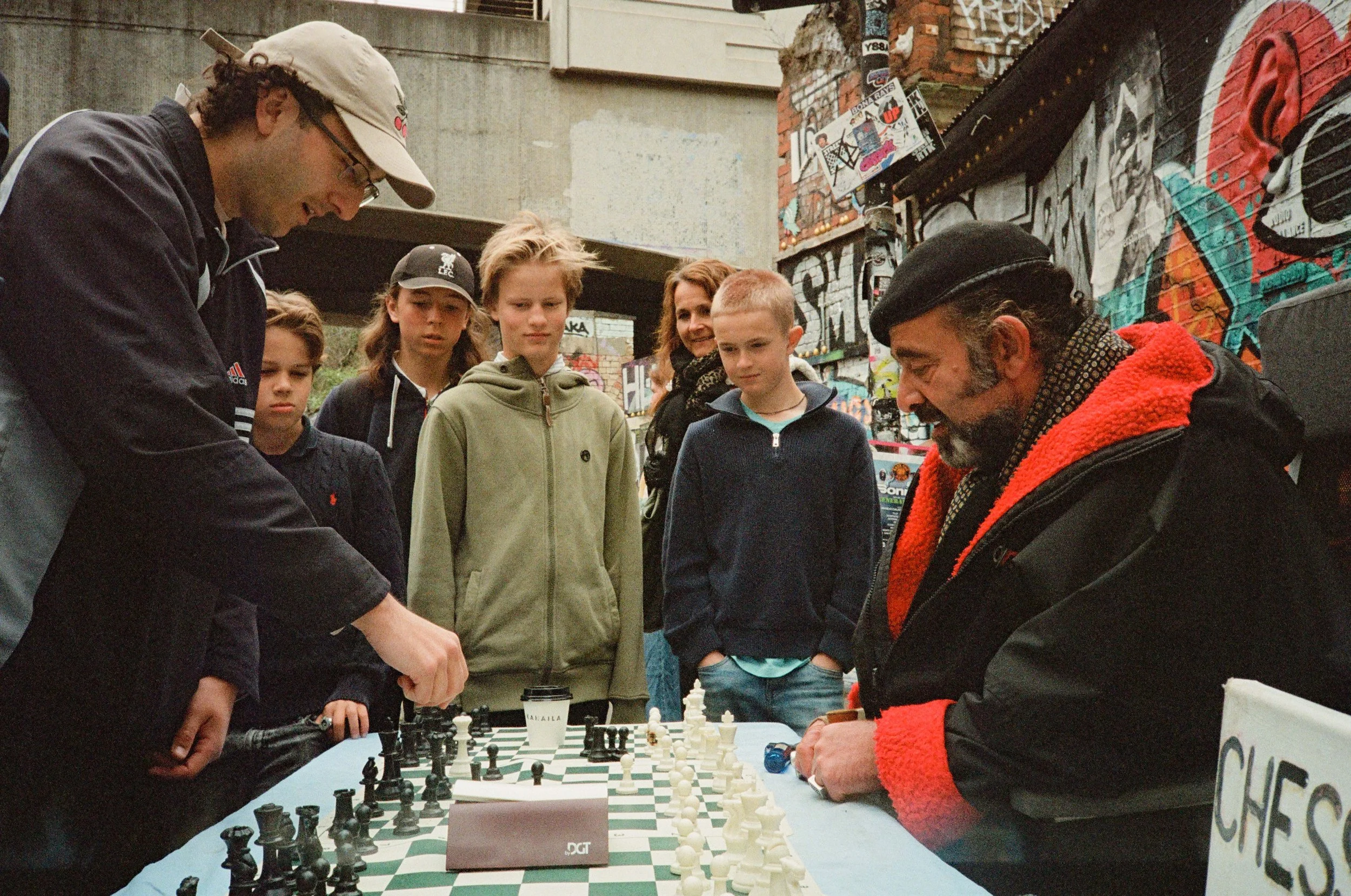 People playing chess at an outdoor event with graffiti-covered walls in the background.
