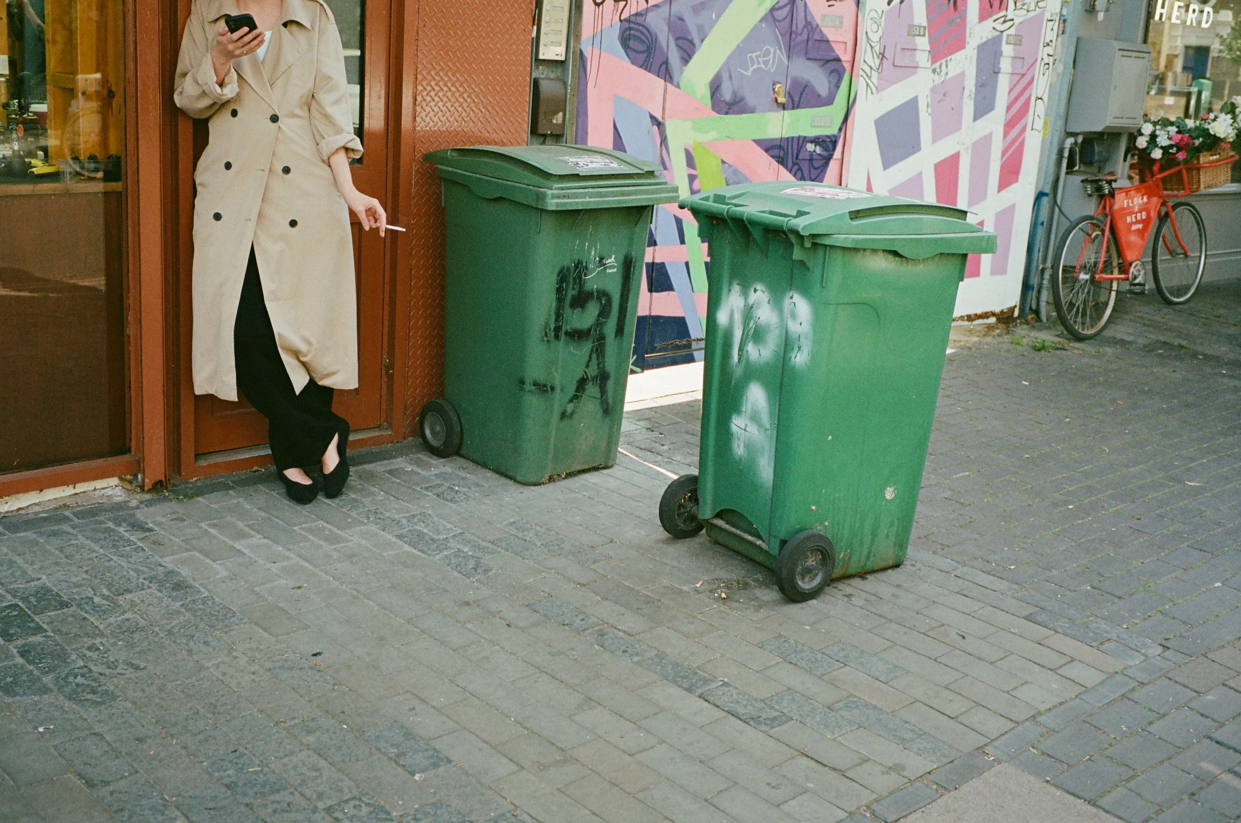 A woman in a beige trench coat is standing on a sidewalk, looking at her phone, with two green trash bins in front of her and a graffiti-covered wall behind. There is also a red bicycle with a flower basket parked nearby.