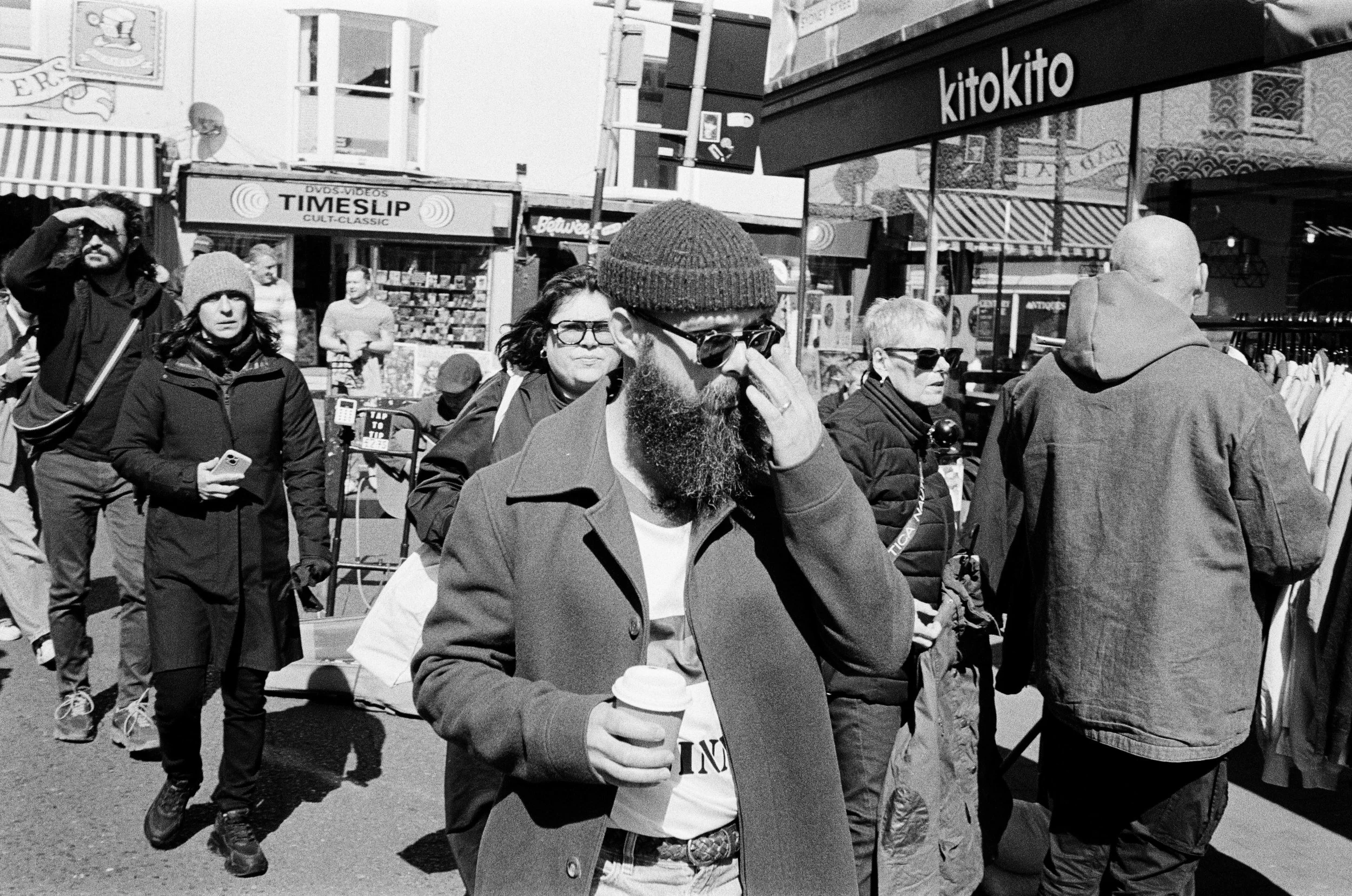 Black and white photo of a group of people walking in a city street. A man in the foreground has a beard, glasses, a beanie, and is holding a coffee cup. Behind him, several women and men are seen walking or standing, some with sunglasses and casual 