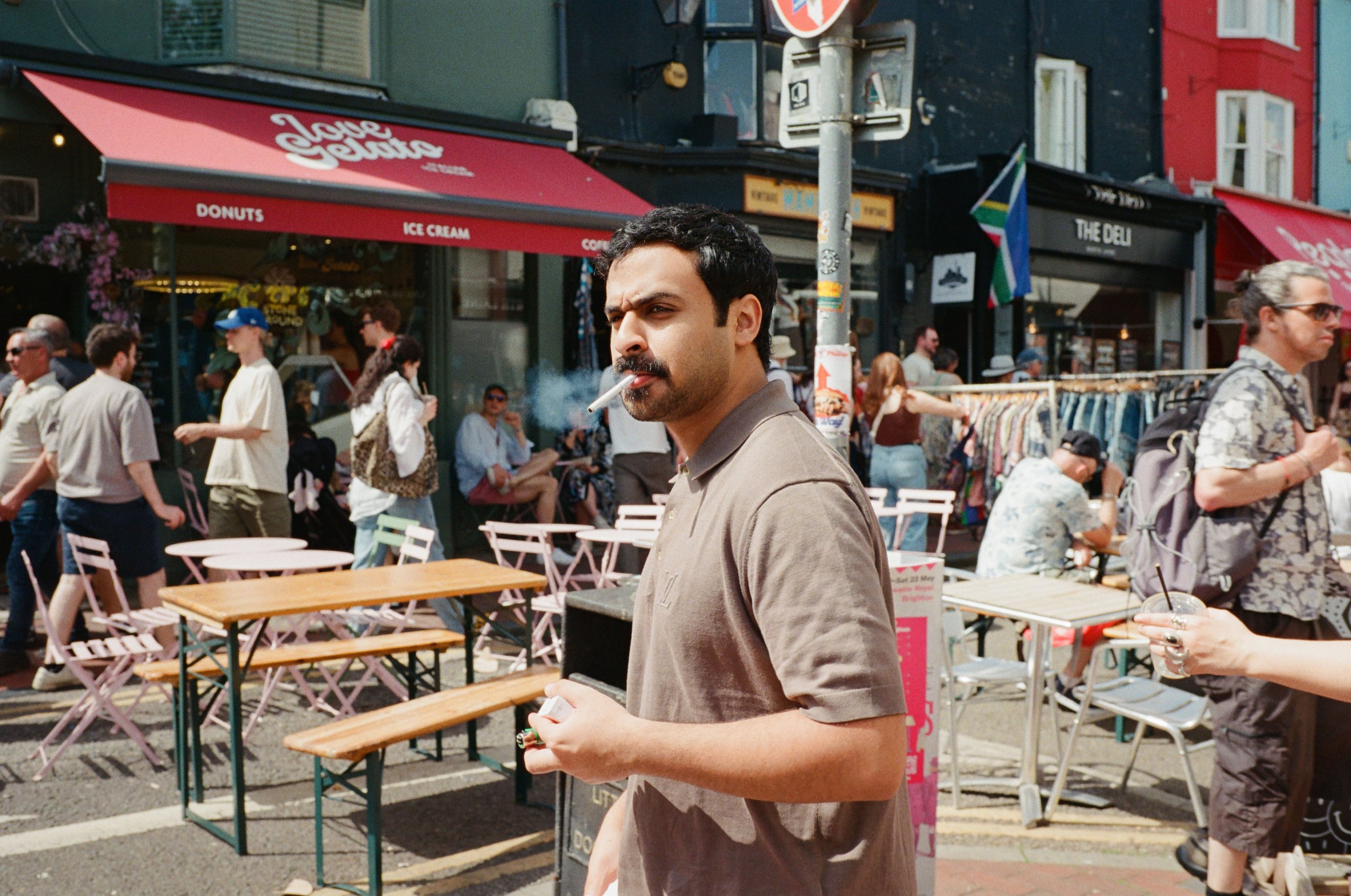 A man with dark hair and a mustache smokes a cigarette while walking on a busy street with outdoor tables, storefronts, and pedestrians.