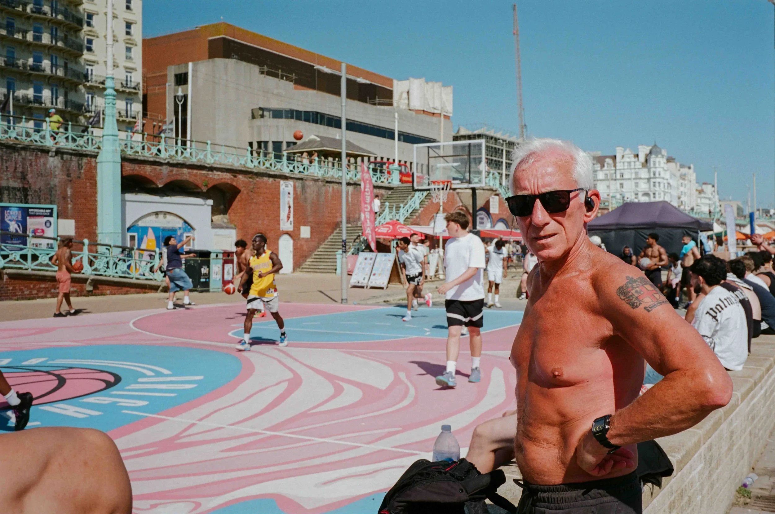 An older shirtless man with white hair, sunglasses, and tattoos on his arm participating in a sunny outdoor basketball game on a colorful court. People are visible in the background, along with buildings and a clear blue sky.