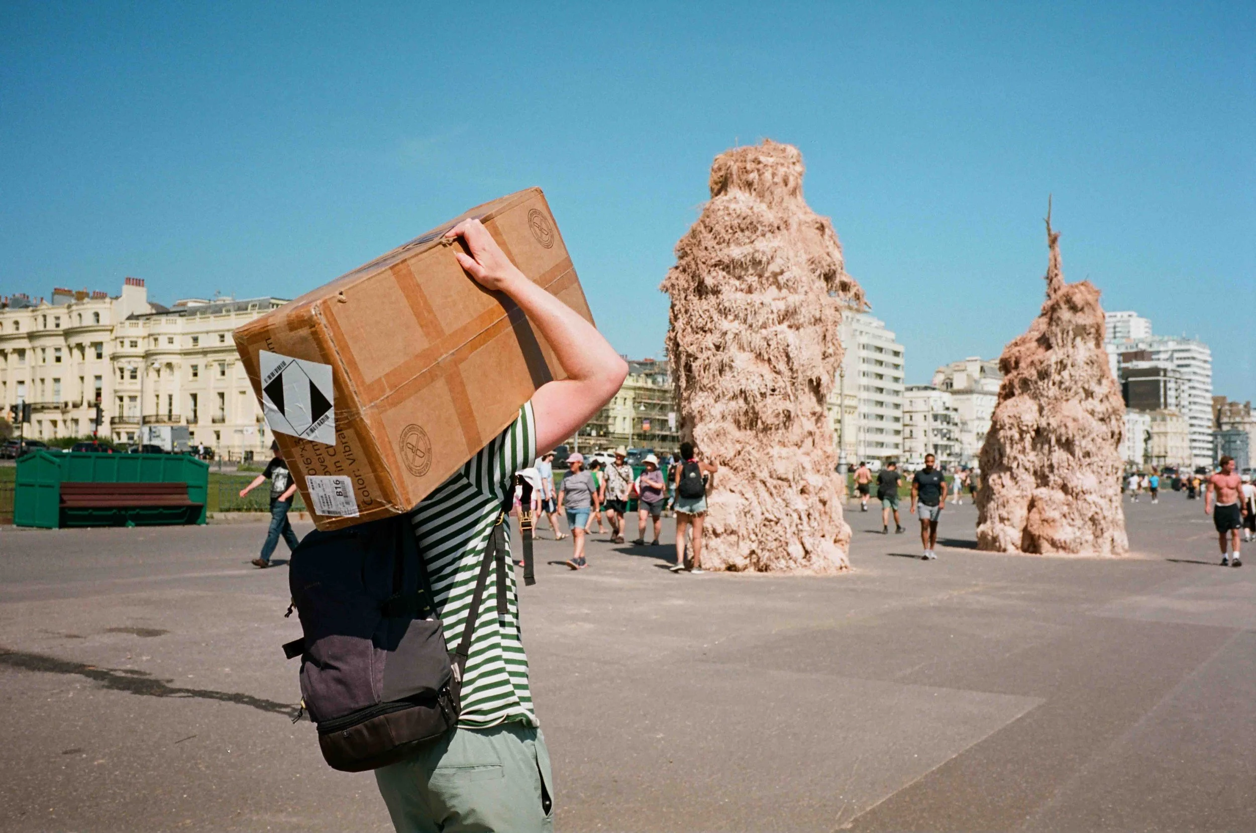 A person carrying a large cardboard box on their shoulder in a busy urban park with sculptures and tall buildings in the background.