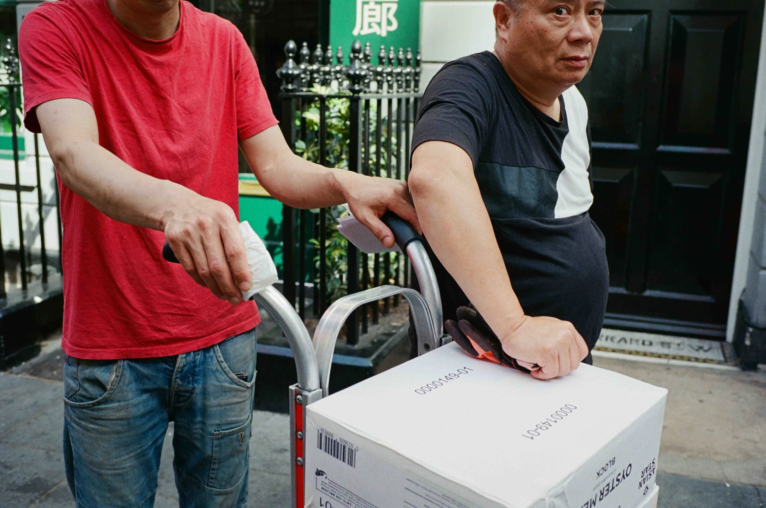 Two men, one wearing a red shirt and the other black, are handling a cardboard box in an outdoor setting. The man in black is in a wheelchair, and the man in red is holding the wheelchair's handle.