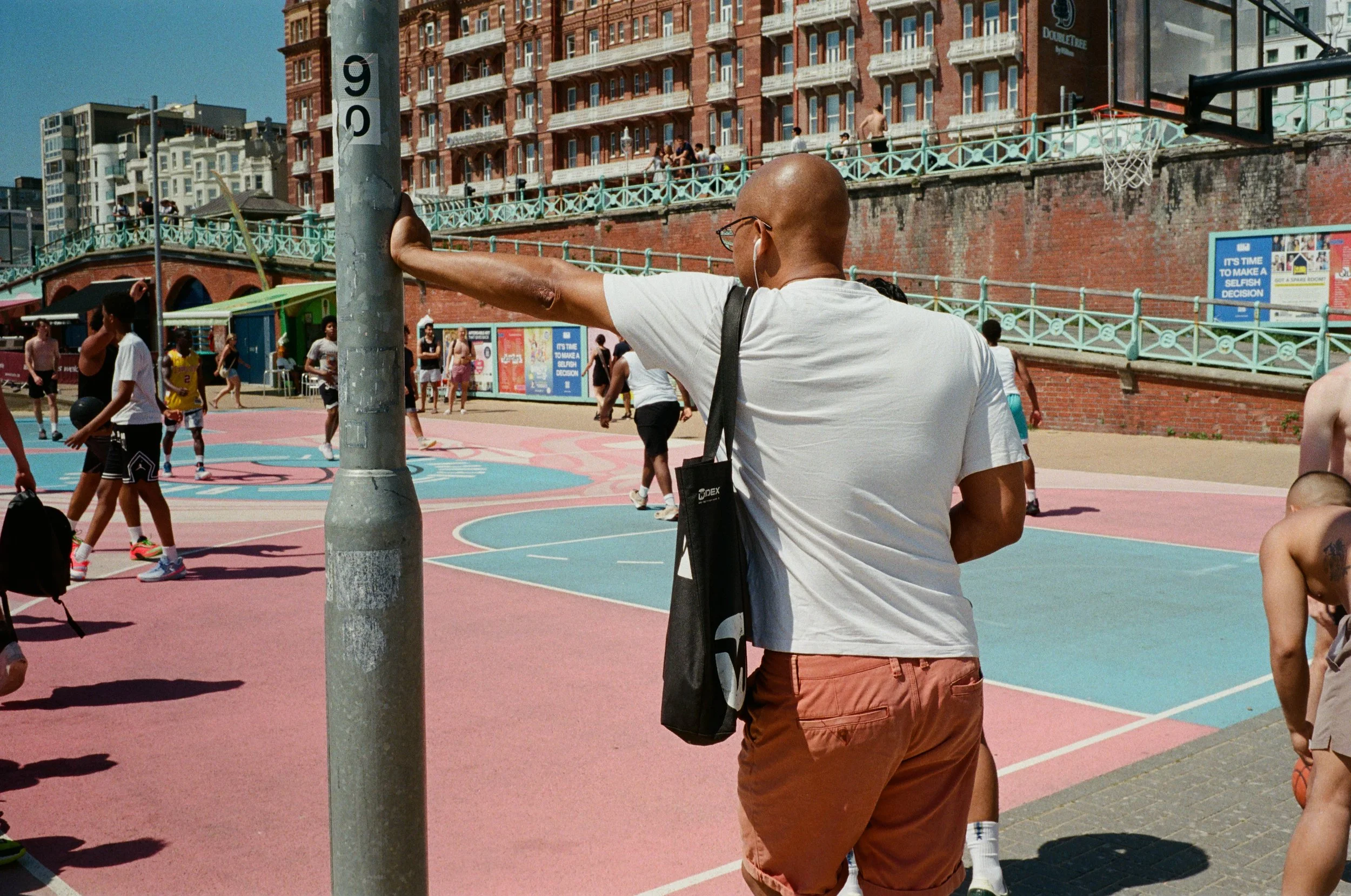 A group of young people playing basketball on an outdoor court in an urban setting. A man with a shaved head, glasses, and carrying a black bag is standing near a basketball hoop, facing away from the camera. Some players are holding basketballs, and
