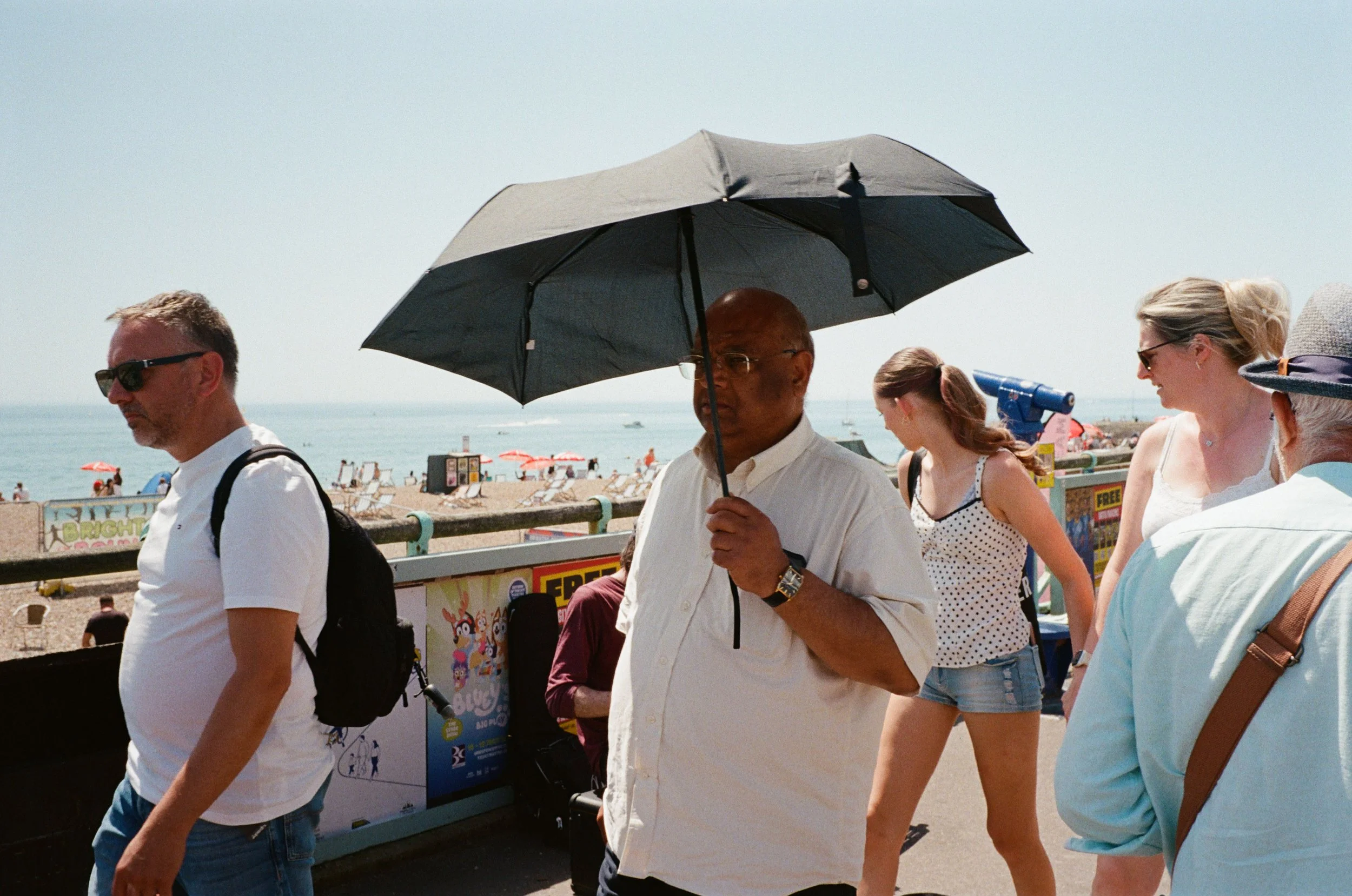 People walking along a beach promenade with umbrellas, beach chairs, and ocean in the background.