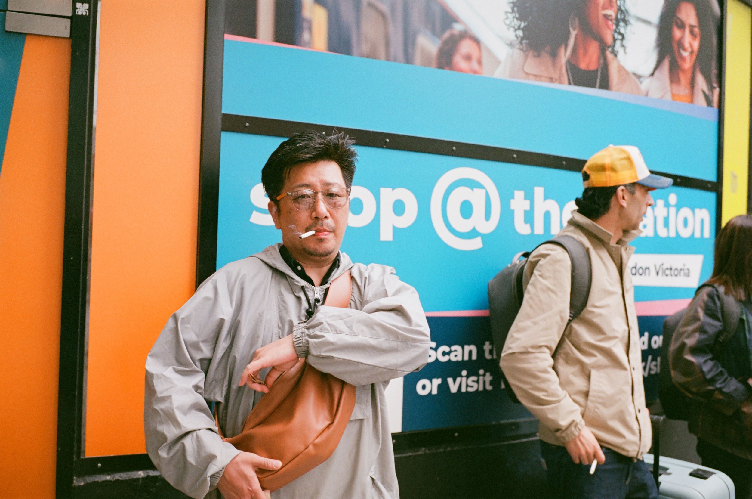 A man with glasses, a gray jacket, and a shoulder bag standing with his arms crossed, smoking a cigarette, and looking at the camera in front of a bright blue advertisement sign with partially visible text.