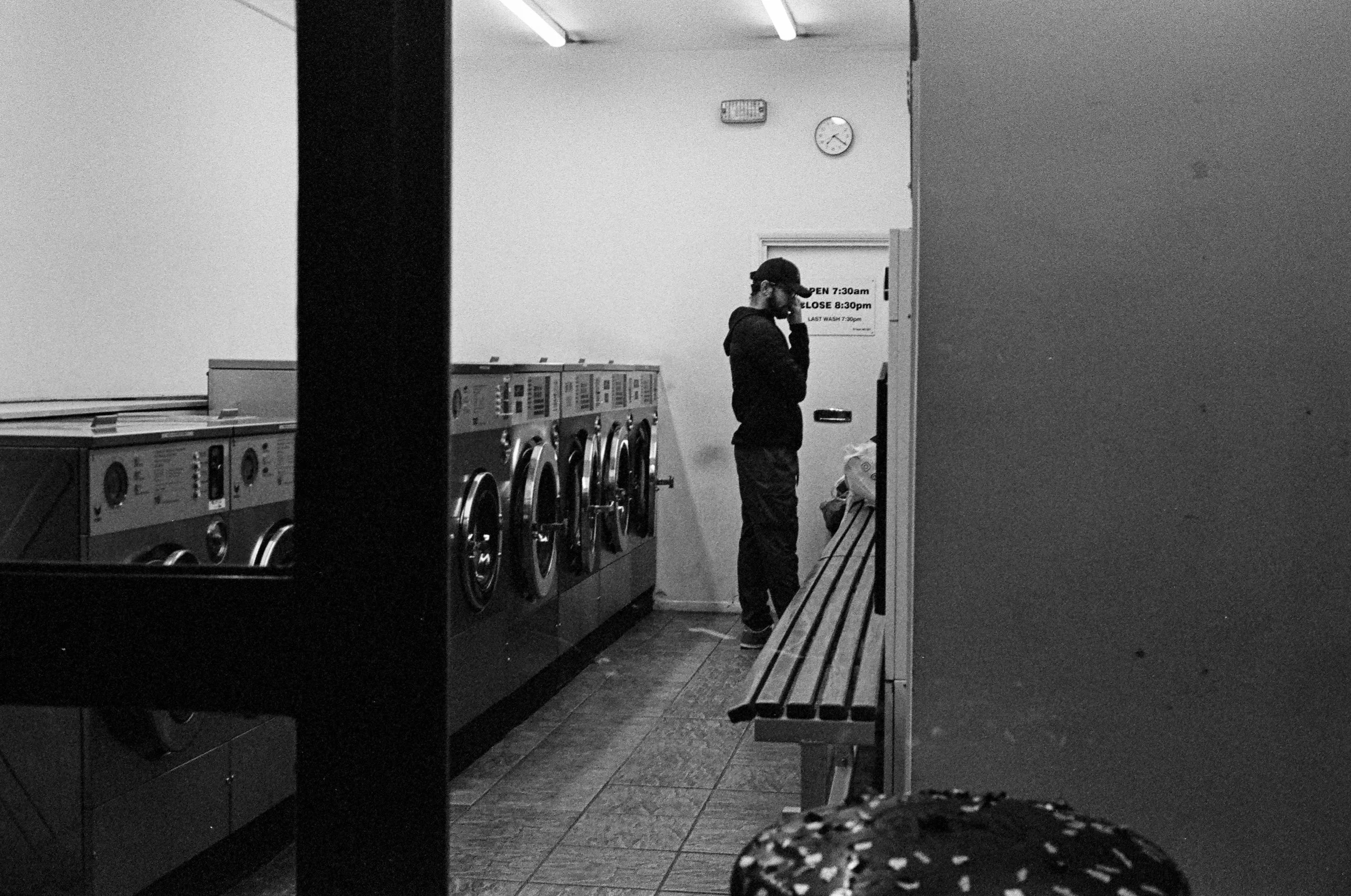A man in a black cap and hoodie stands inside a laundry room, looking at a machine. There are multiple washing machines lined up, a bench, and a sign on the wall indicating opening and closing times. The room has a clock showing 2:55.