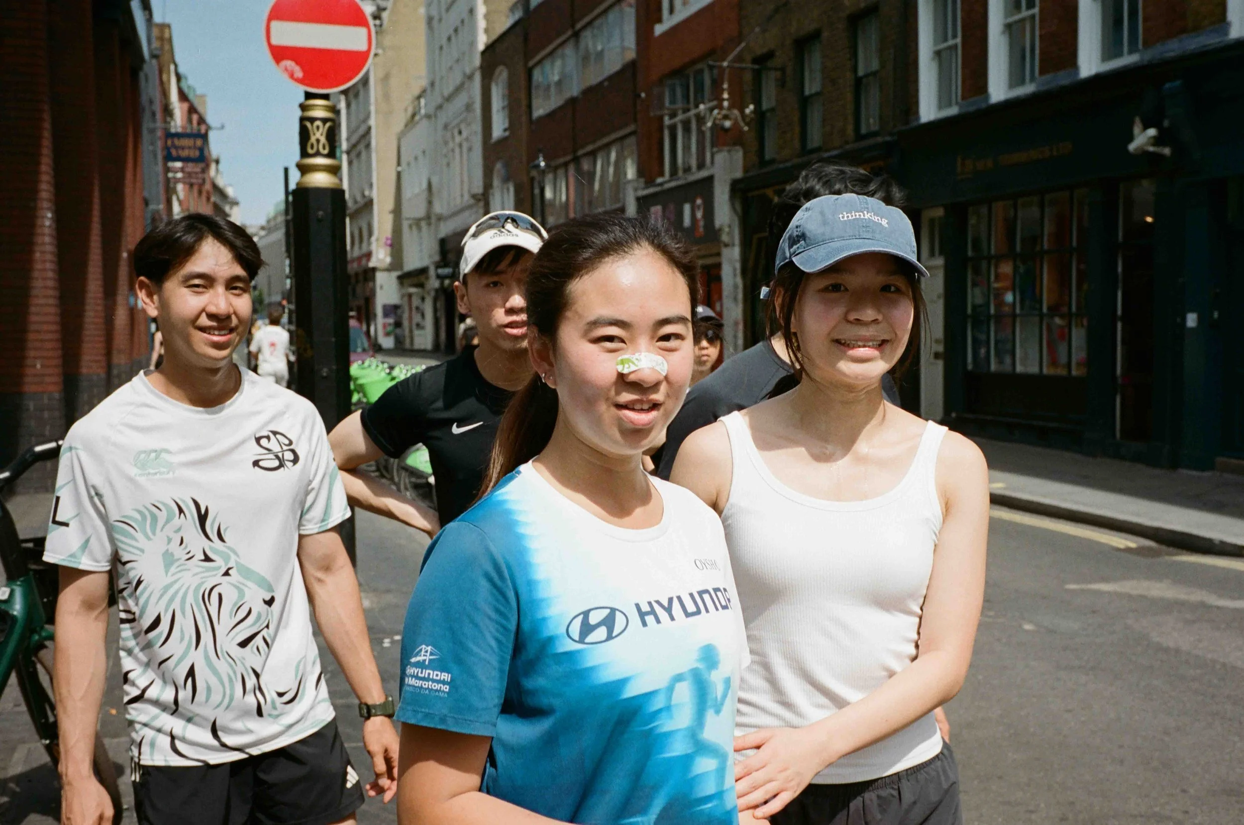 A group of runners standing together on a city street during a race, smiling and looking at the camera, with buildings and shops in the background.