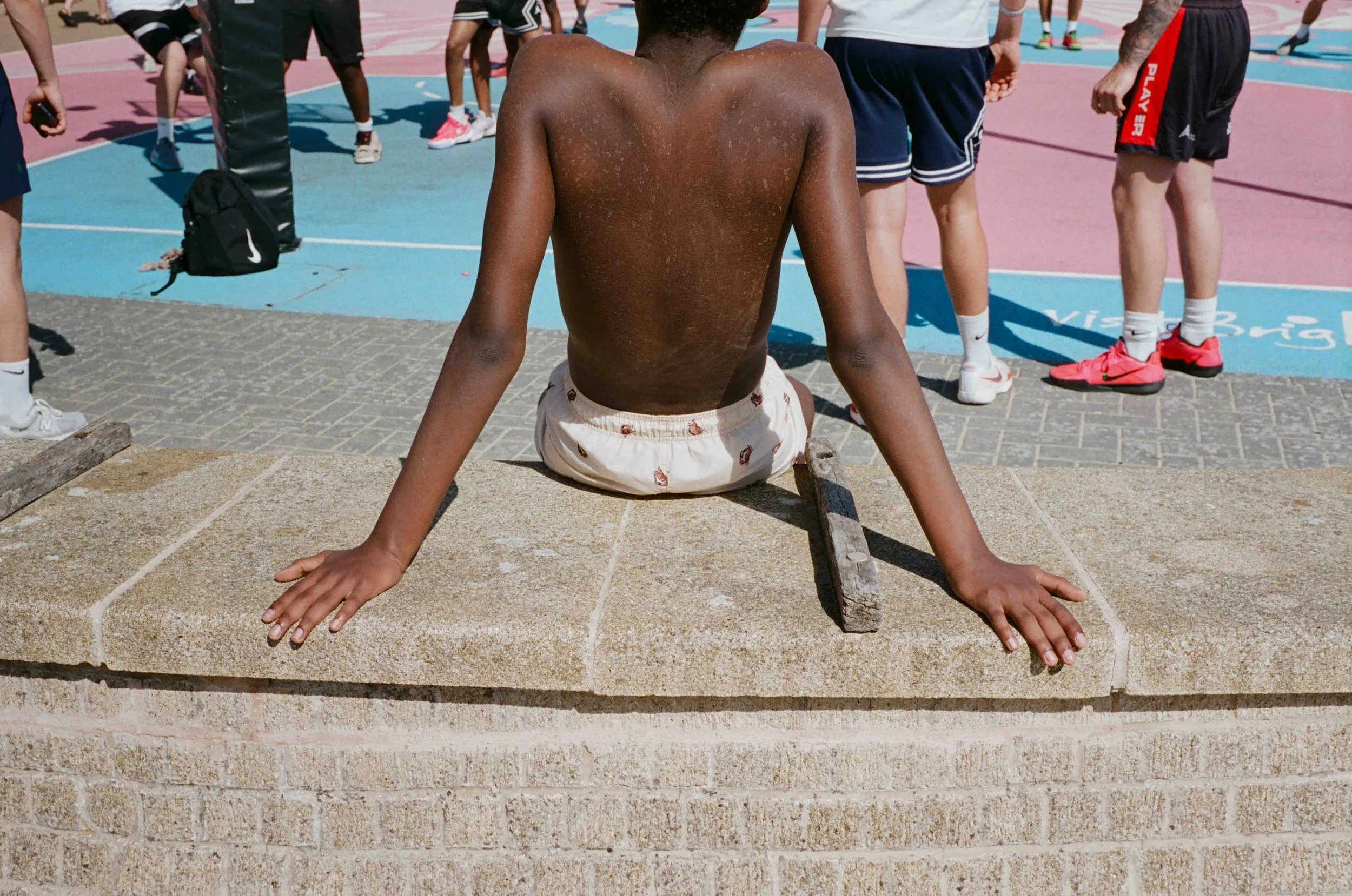 A young boy sitting on a sidewalk with his back facing the camera, surrounded by other children and adults near a colorful outdoor basketball court.