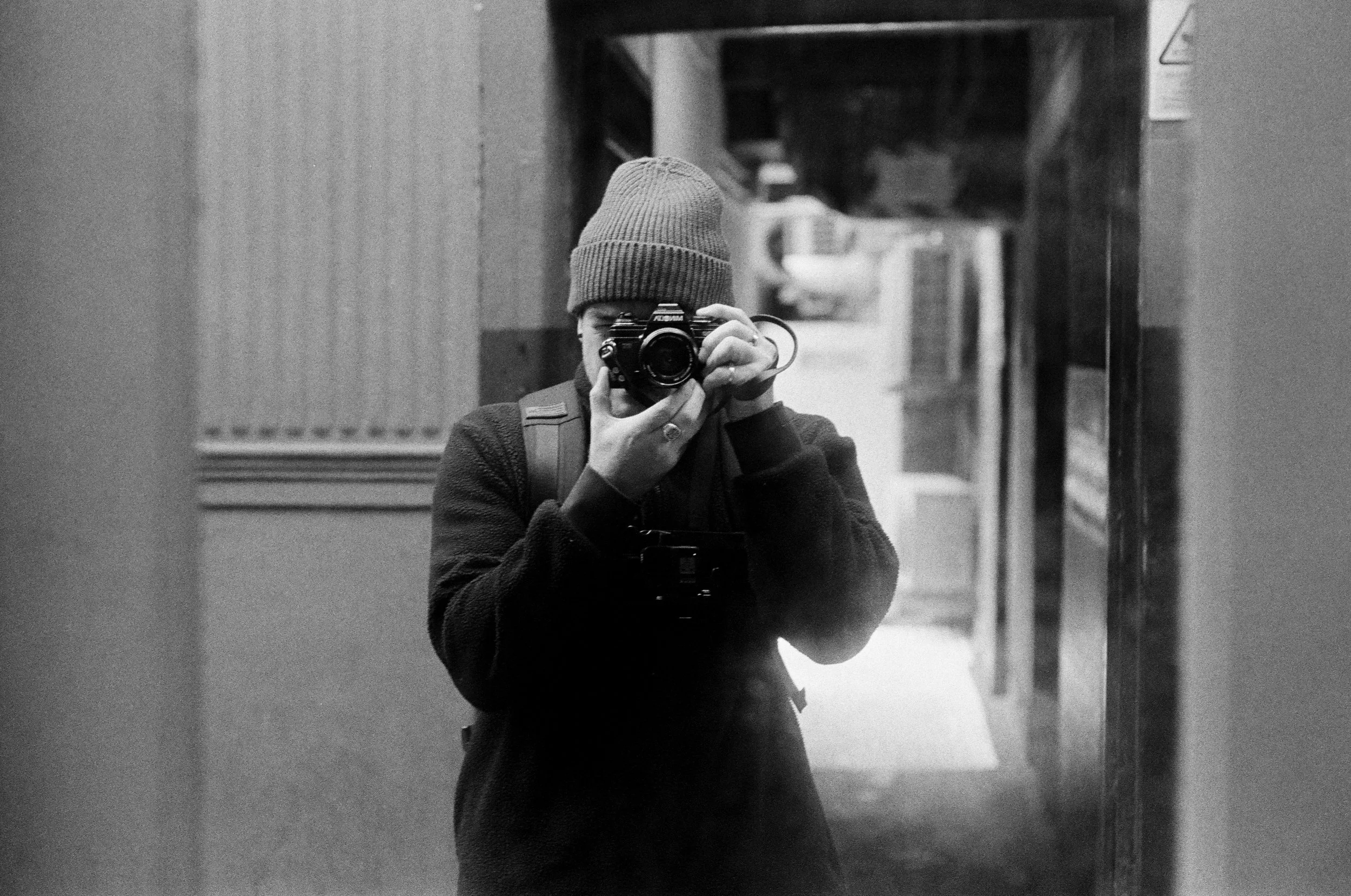 A person wearing a beanie and dark jacket taking a photo of themselves in a mirror, holding a camera up to their face.