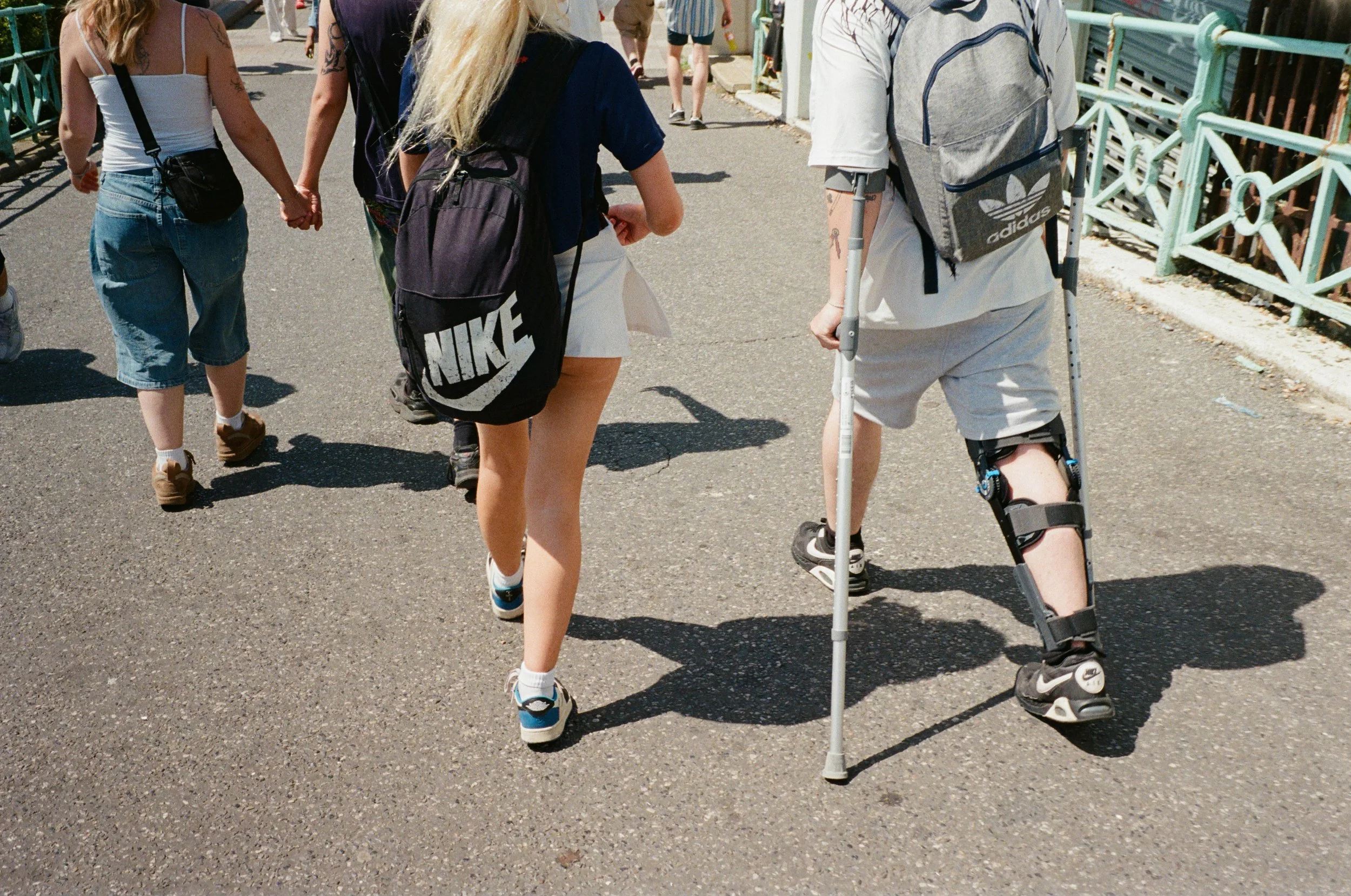 A group of people walking on a sidewalk, including a woman with long blonde hair, a woman with a gray backpack with Adidas logo, and a person with a leg brace on crutches wearing sneakers.