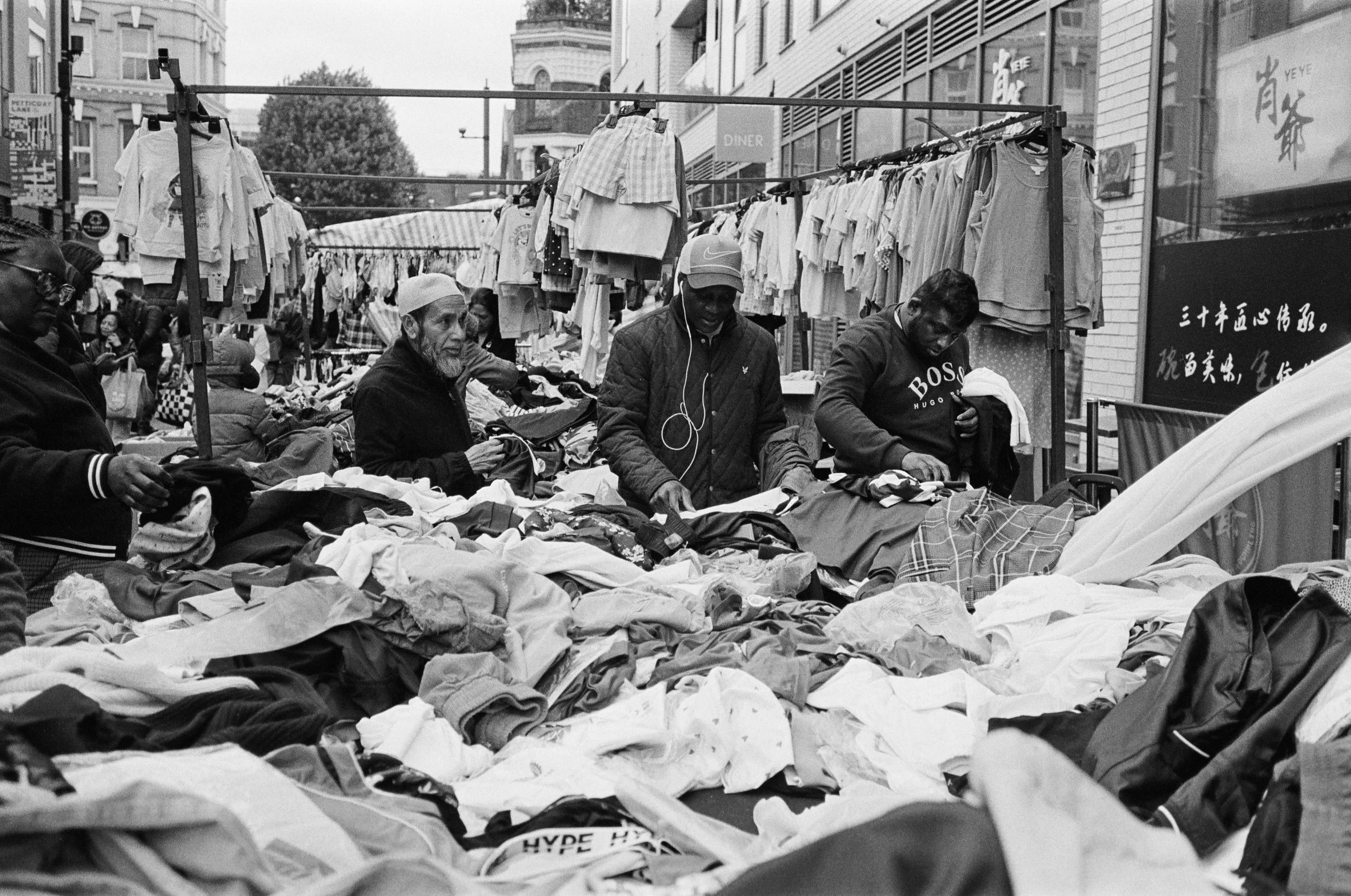 Street market scene with vendors selling clothes on tables and hanging on racks amid city buildings.