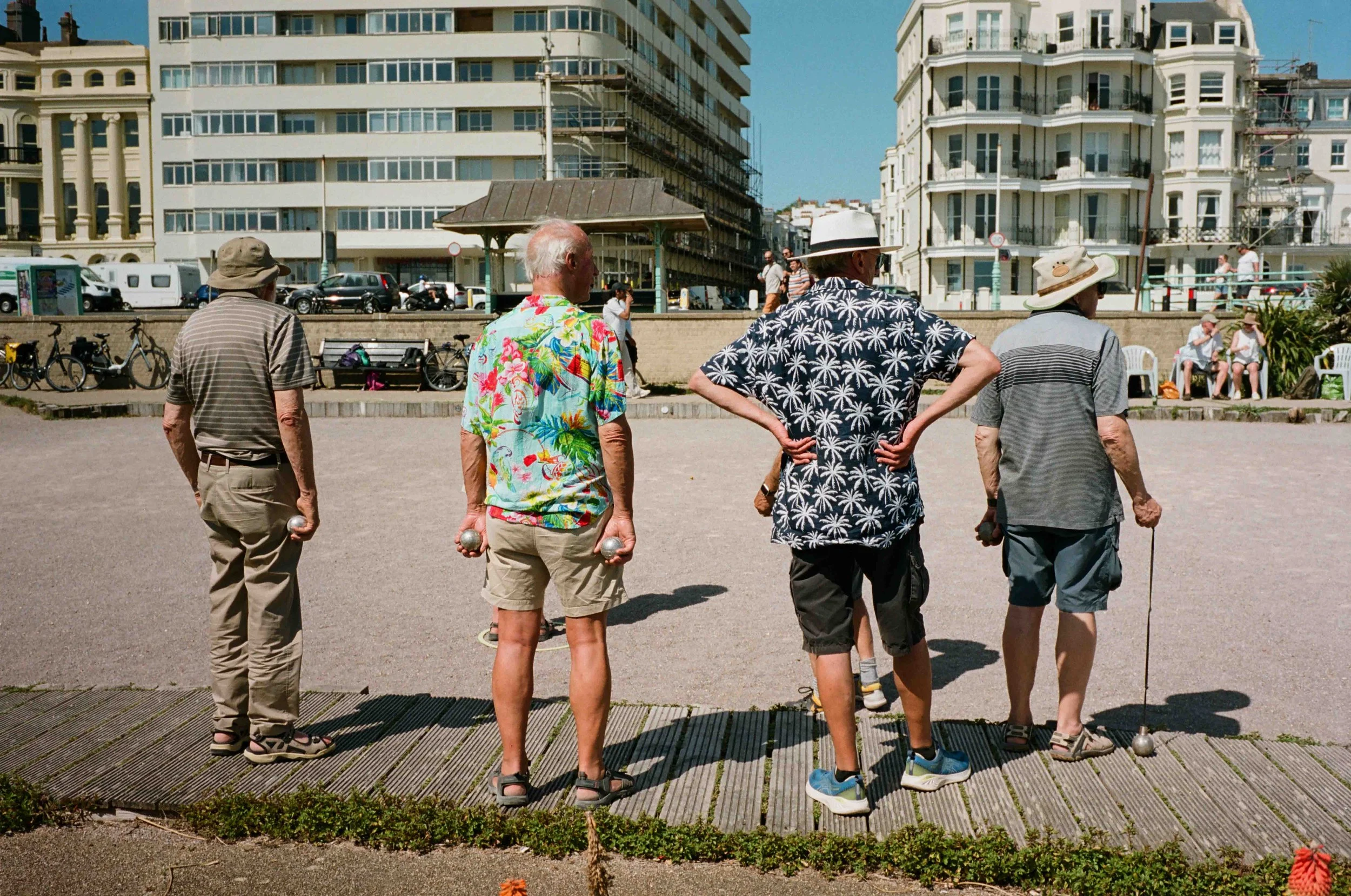 Older men playing pétanque outdoors in an urban setting with multi-story buildings and onlookers in the background.