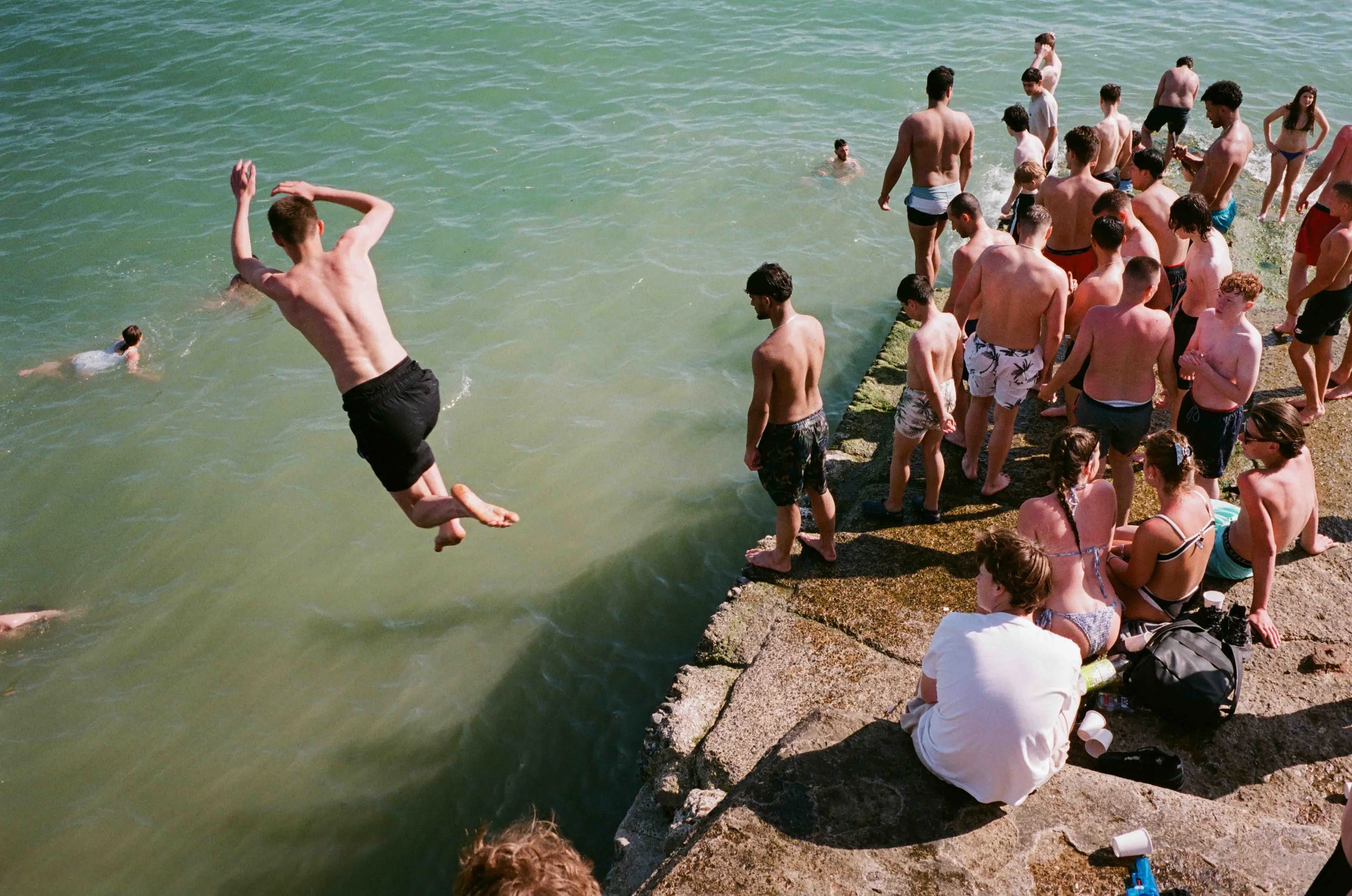 People at a beach, some swimming in the water, others standing or sitting on the rocky shore, with one person mid-air jumping into the water.
