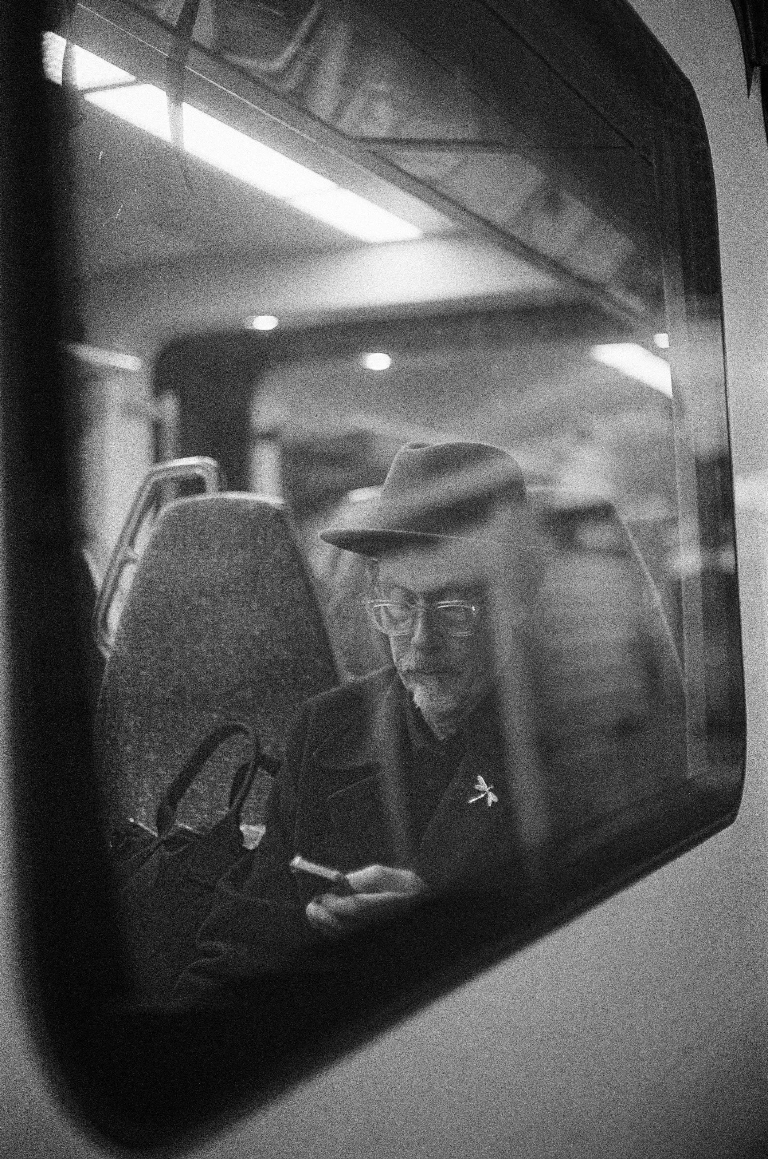 A man with glasses and a hat sitting on a bus or train, looking at his phone, seen through a window.