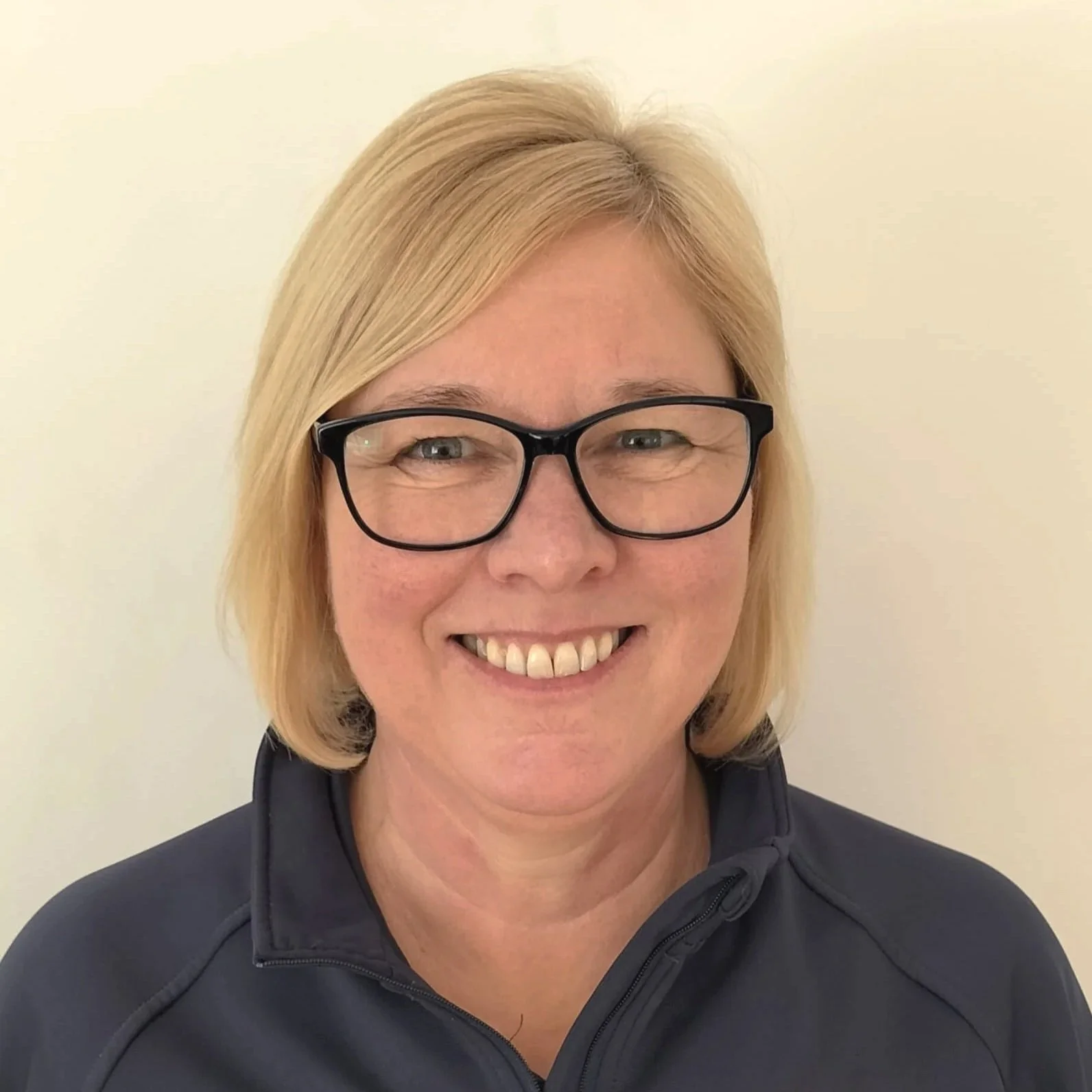 Close-up photo of a smiling woman with blonde hair, wearing black glasses and a dark zip-up jacket, standing against a plain light-colored background.