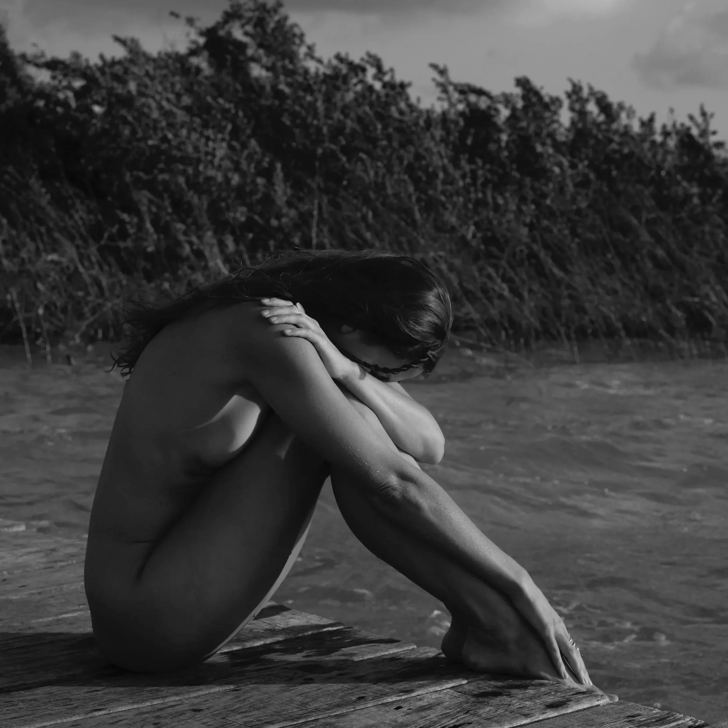 A black and white photo of a nude woman sitting on a wooden dock by the water, hugging her knees and resting her forehead on her arms, with a natural landscape and cloudy sky in the background.