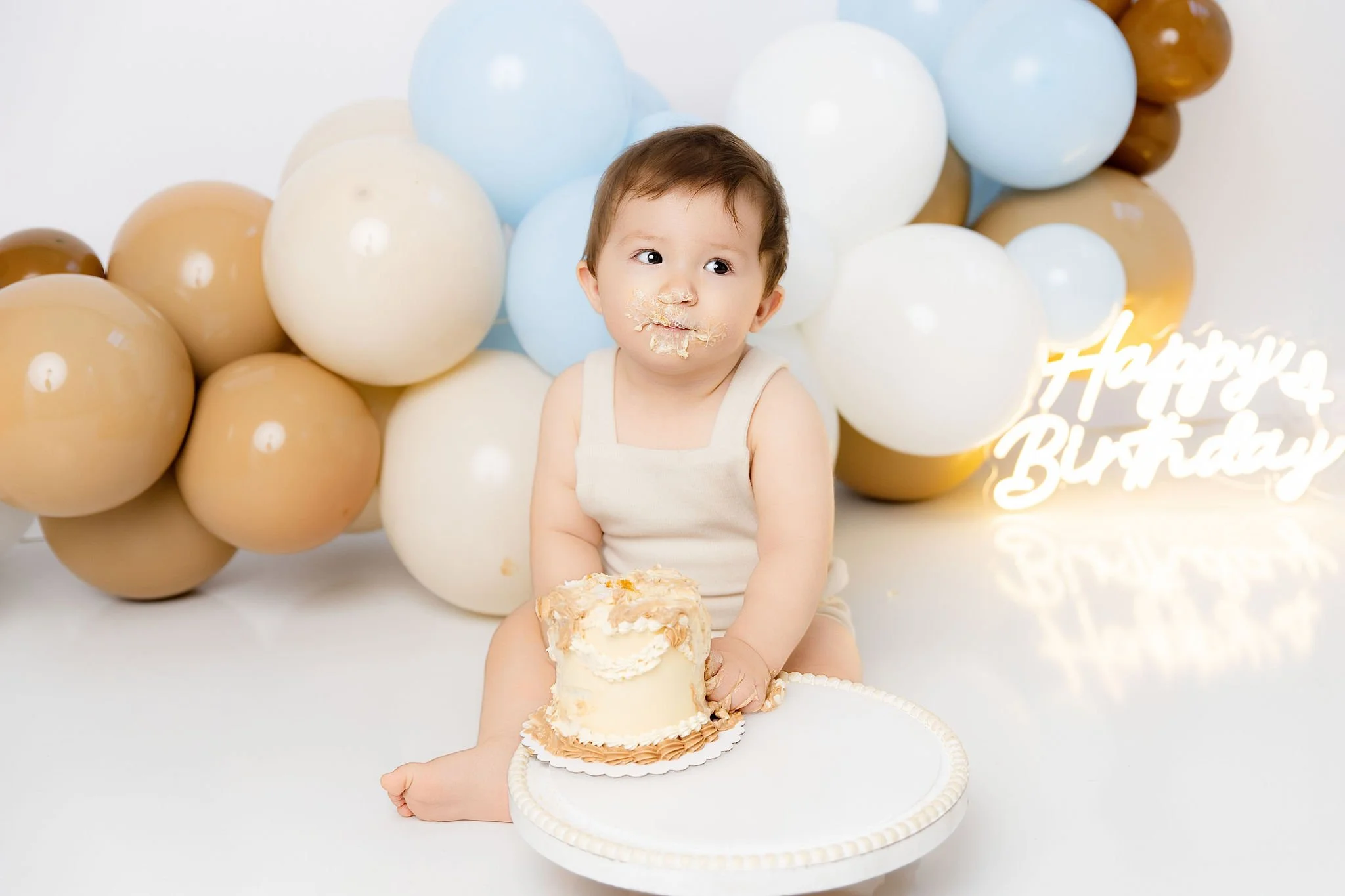A young child with cake and frosting on their face and hands, sitting on the floor with a small cake on a white tray, in front of a backdrop of cream, tan, light blue, and white balloons and a 'Happy Birthday' neon sign.