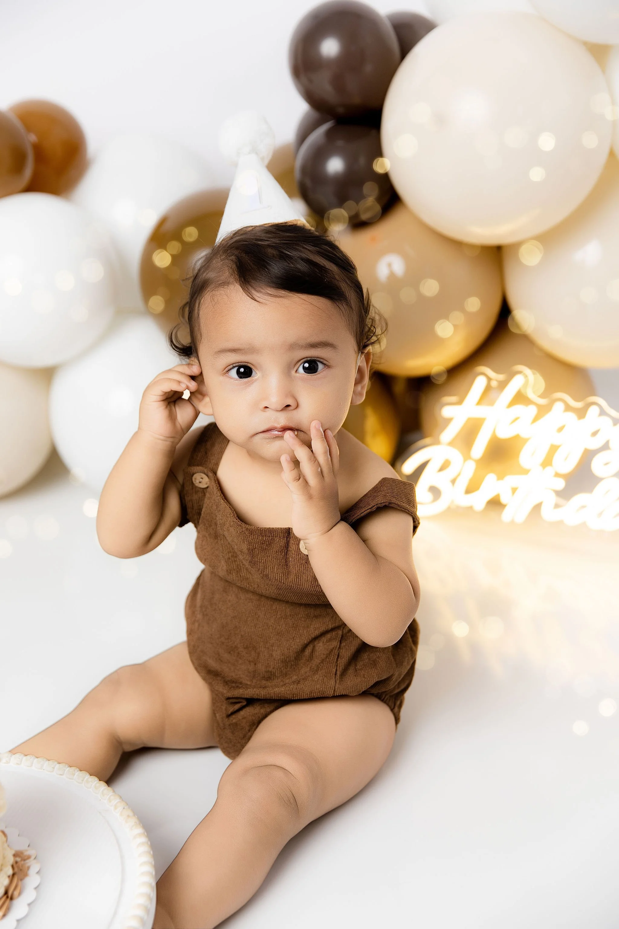 Baby sitting on the floor at birthday celebration with brown, white, and gold balloons in the background and a glowing sign that says "Happy Birthday."