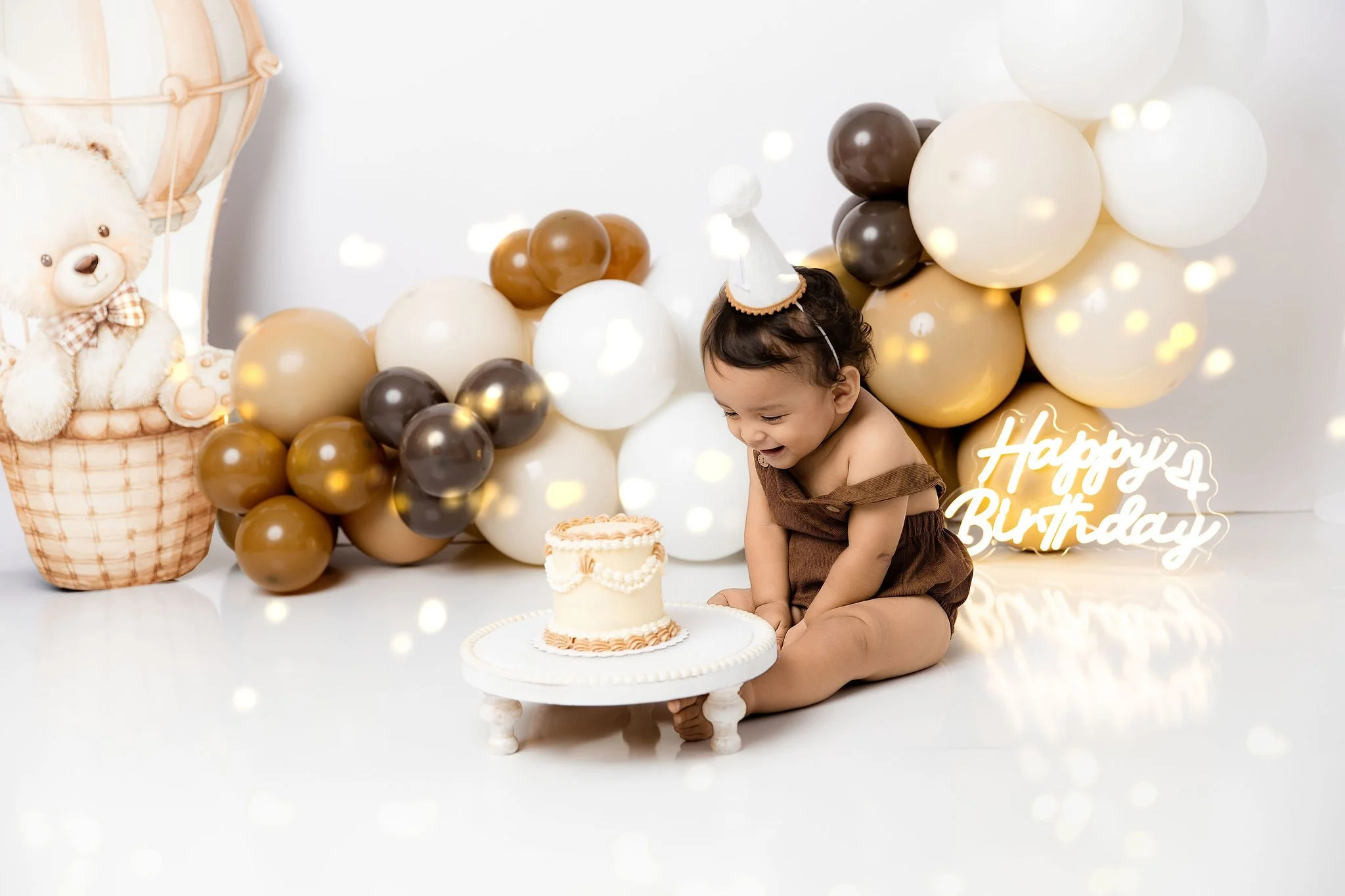 Baby celebrating birthday with a small cake, wearing a party hat, surrounded by balloons and a teddy bear plush in a decorated setting.