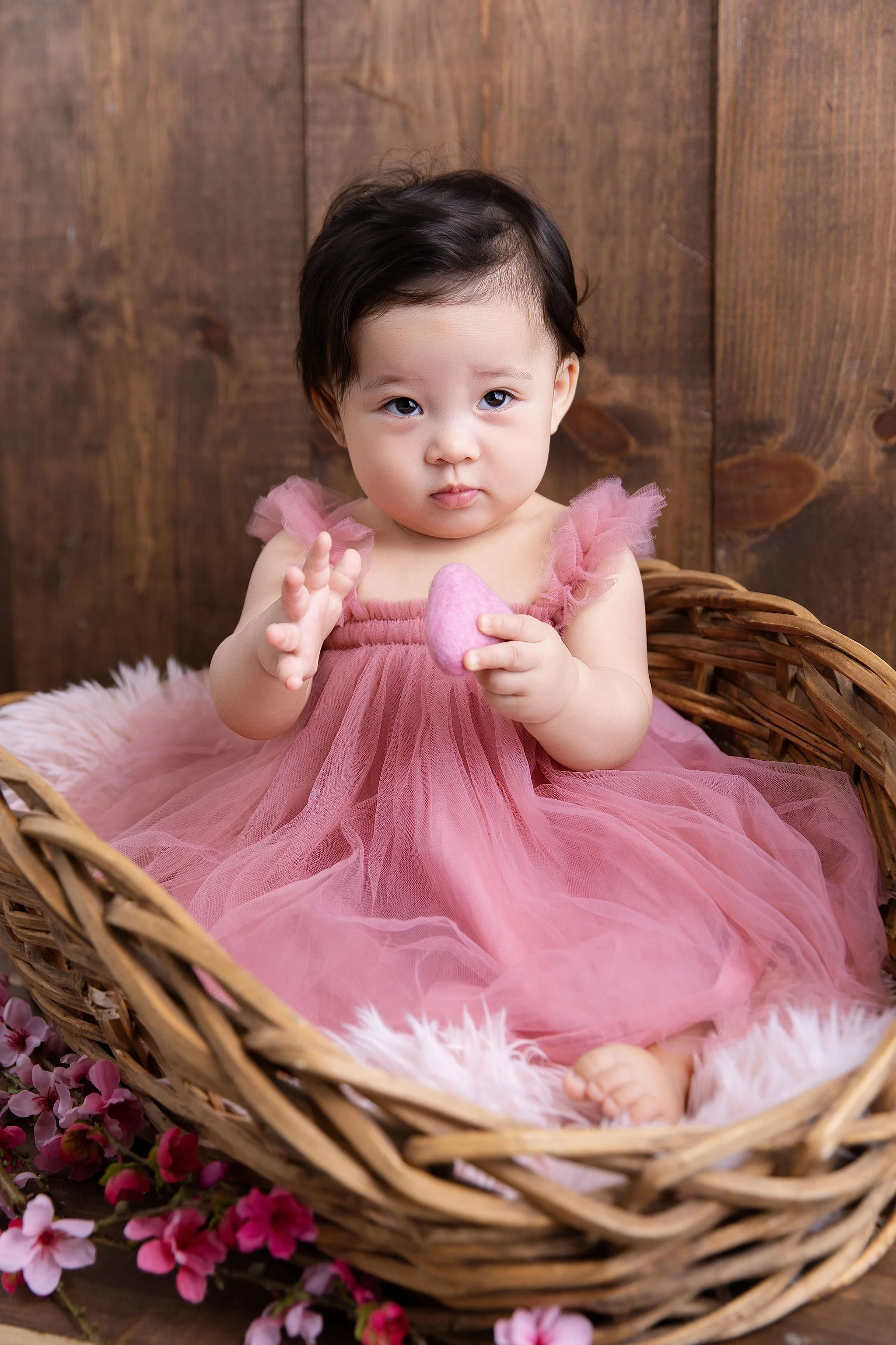 A young girl in a pink tulle dress sitting in a wicker basket surrounded by pink flowers and holding a pink heart-shaped object.