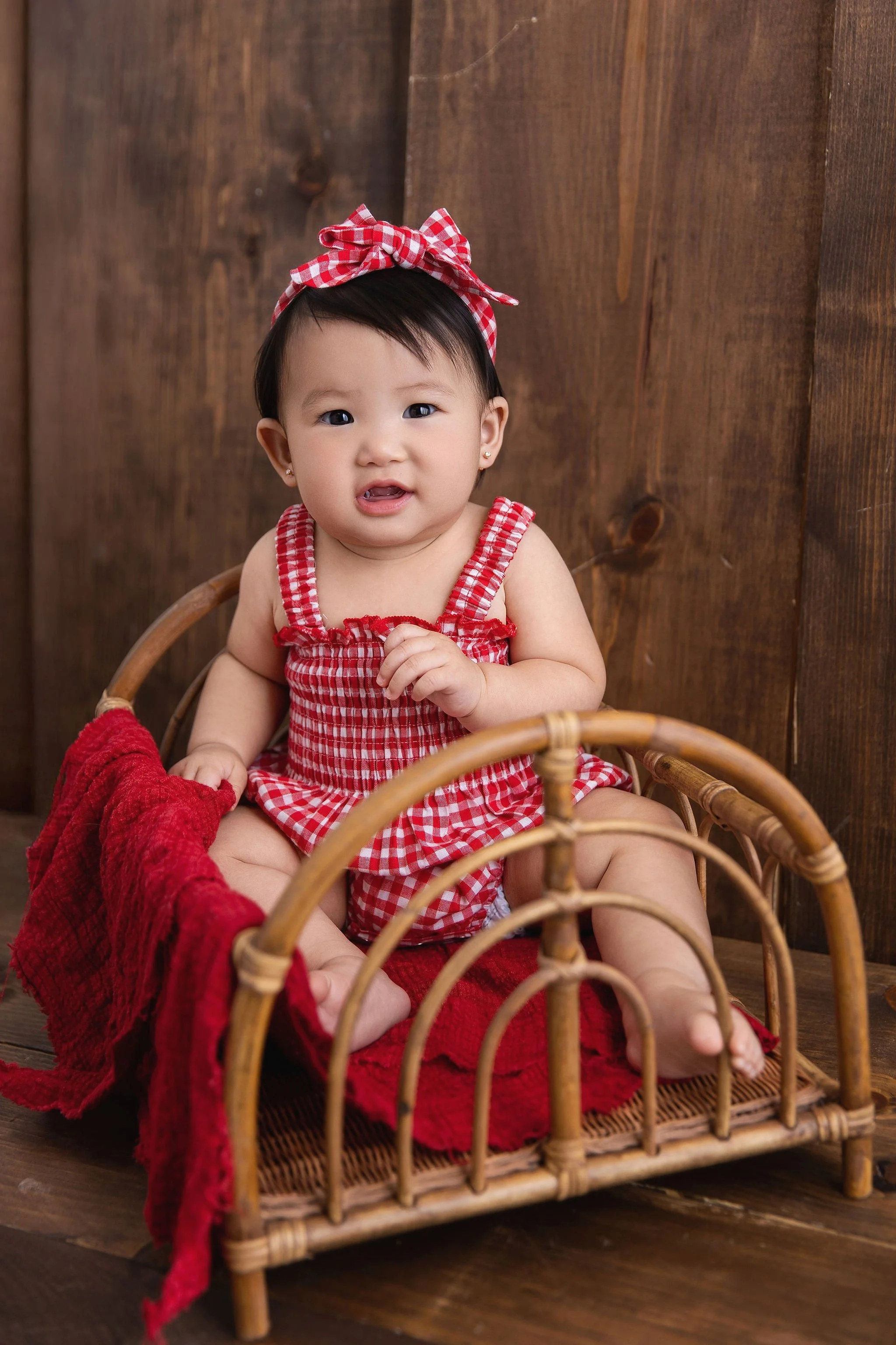 Young girl with short black hair, wearing a red and white checkered outfit and matching headband, sitting on a small wicker bed with a red blanket, against a wooden wall background.