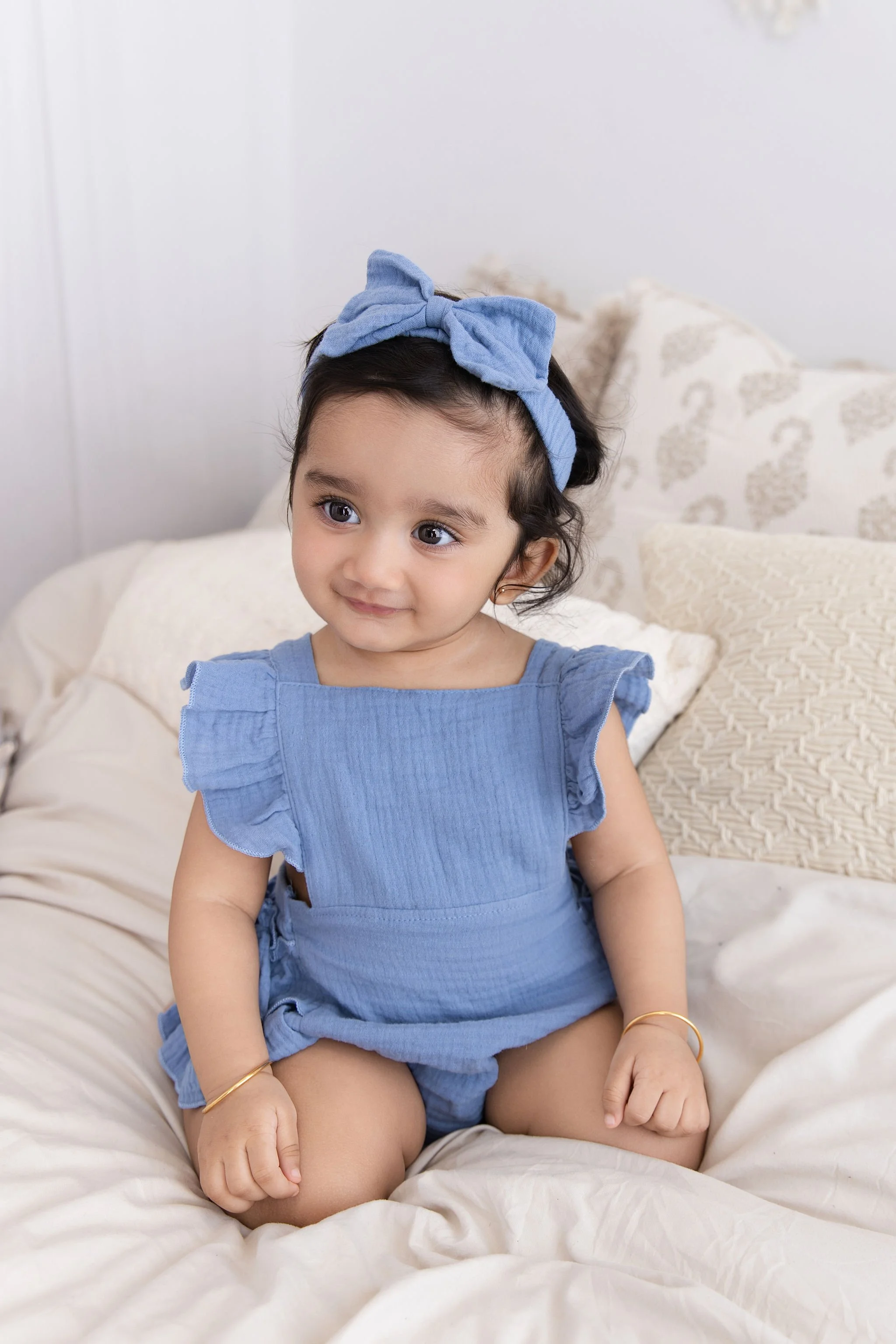 A young girl with dark hair and big eyes, wearing a blue dress and matching headband, sitting on a bed with white bedding and patterned pillows.