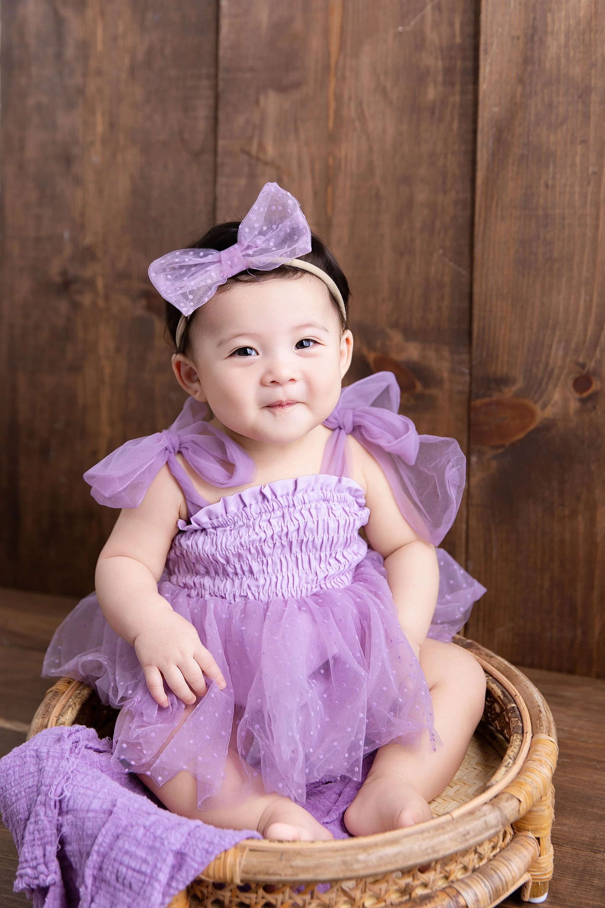 A young girl in a purple dress with tulle fabric, sitting in a wicker basket on a wooden floor with a wooden wall behind her.