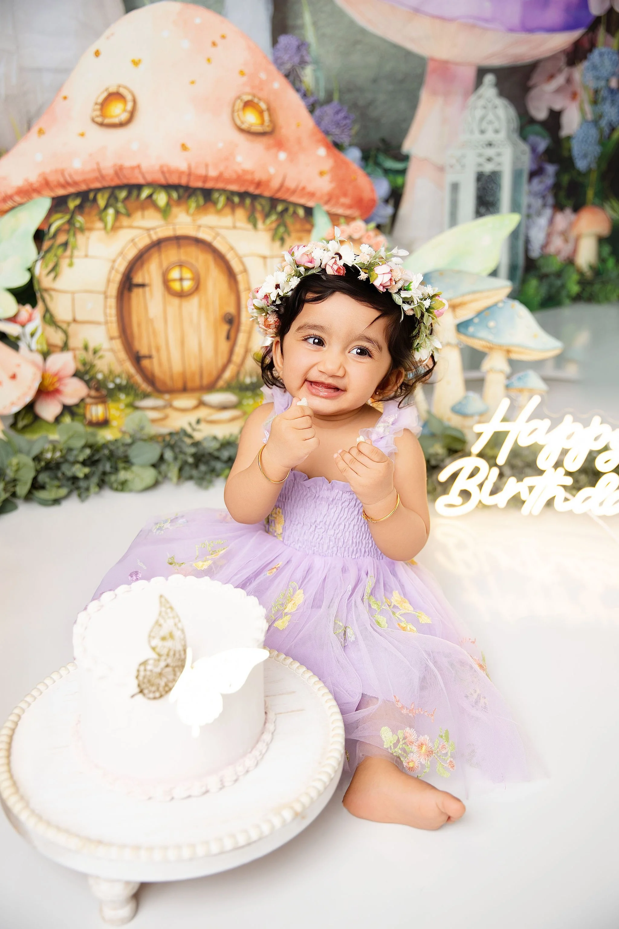 A young girl dressed in a purple tulle dress with floral embroidery, wearing a flower crown, sitting on a white surface with a small white birthday cake decorated with a butterfly topper in front of her. The background features whimsical fairy tale m