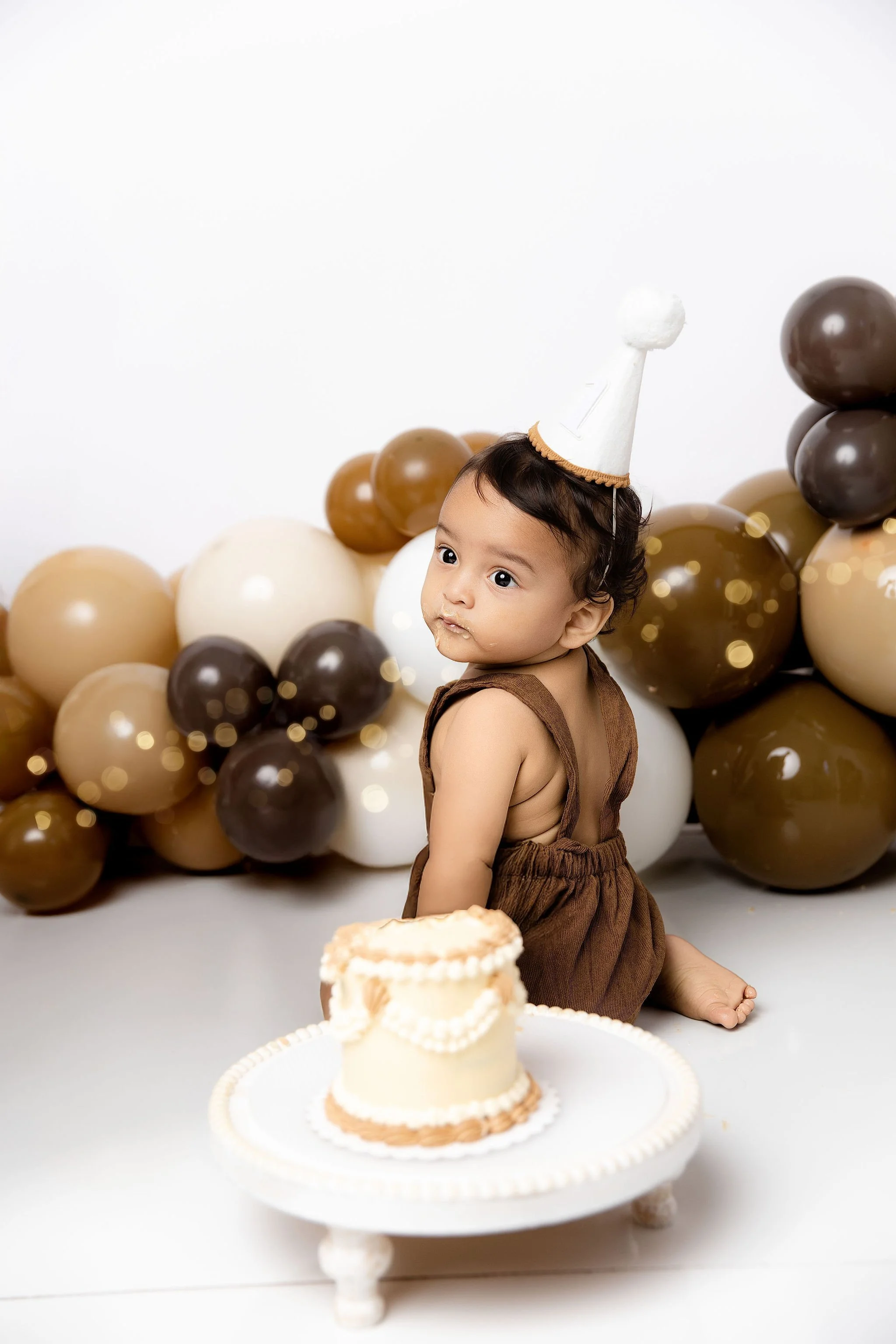 Baby wearing brown romper, sitting on white floor after cake smash, with chocolate and beige balloons in the background, and a small cake on a white stand in front.