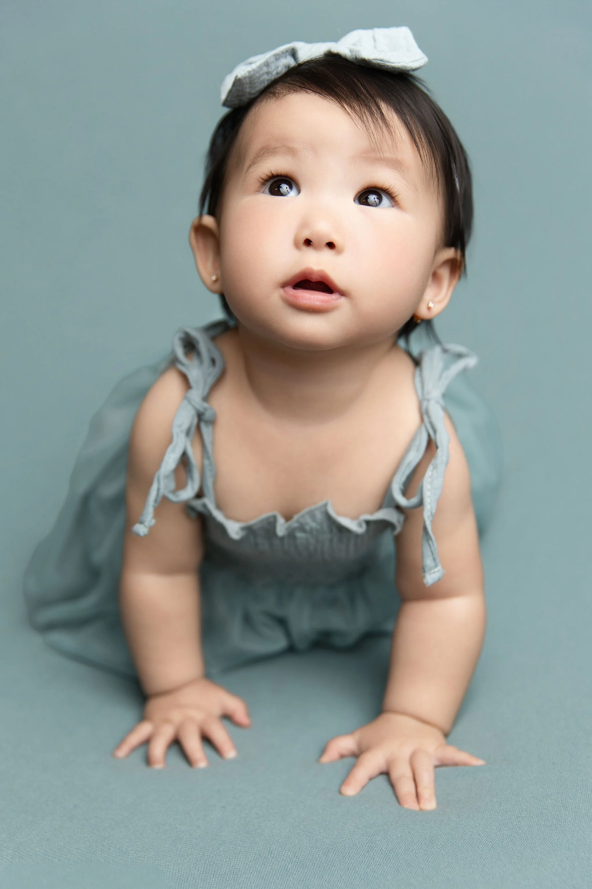 A young girl with dark hair, wearing a gray dress with tied shoulder straps and a large gray bow on her head, poses on her hands and knees looking upwards.
