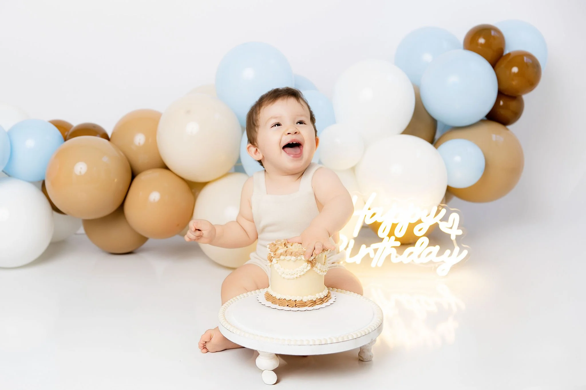 A happy young child sitting in front of a white cake on a cake stand, celebrating a birthday with a smiling face, surrounded by beige, white, and blue balloons and a neon sign that says 'Happy 4th Birthday'.