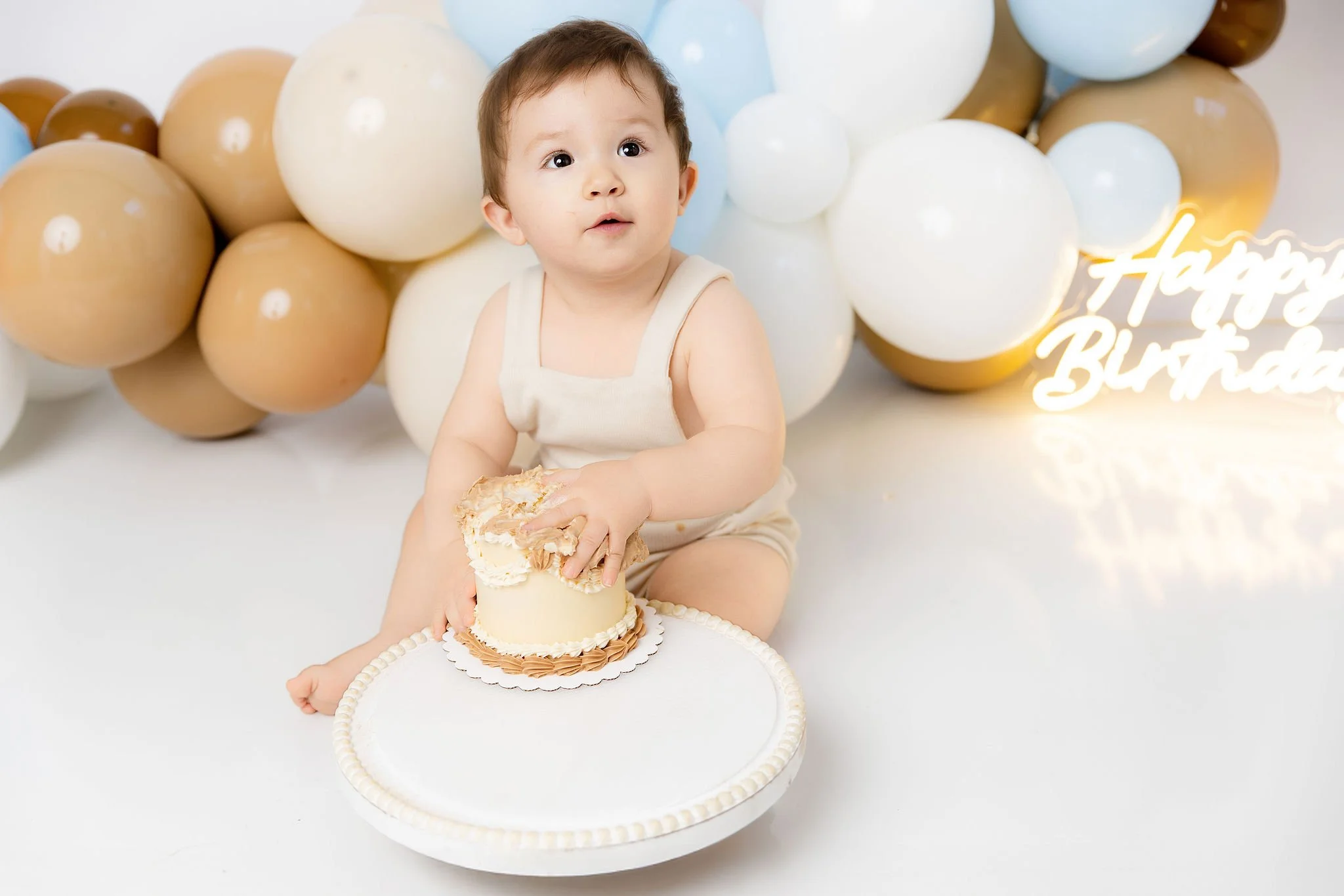 A young boy sitting in front of a tan, white, and light blue balloon backdrop, holding a cake with cream and frosting. A neon sign that says 'Happy Birthday' is visible on the right side of the image.