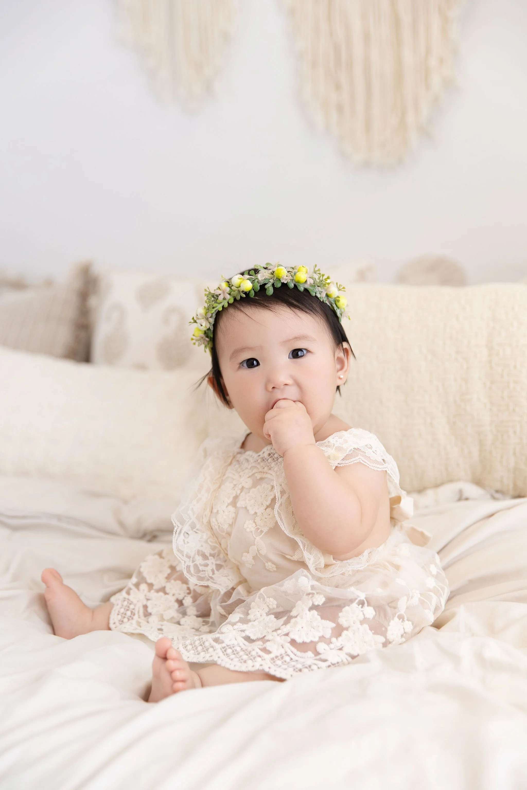 A baby girl with dark hair, dressed in a lace dress, sitting on a bed with cream-colored linens and pillows, wearing a floral crown, and sucking on her fingers.
